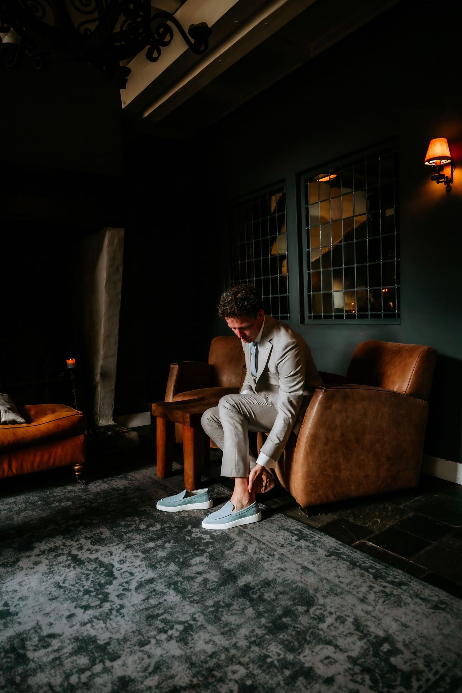 A person in a light colored suit sits alone in a dimly lit lounge with leather furniture and green walls.