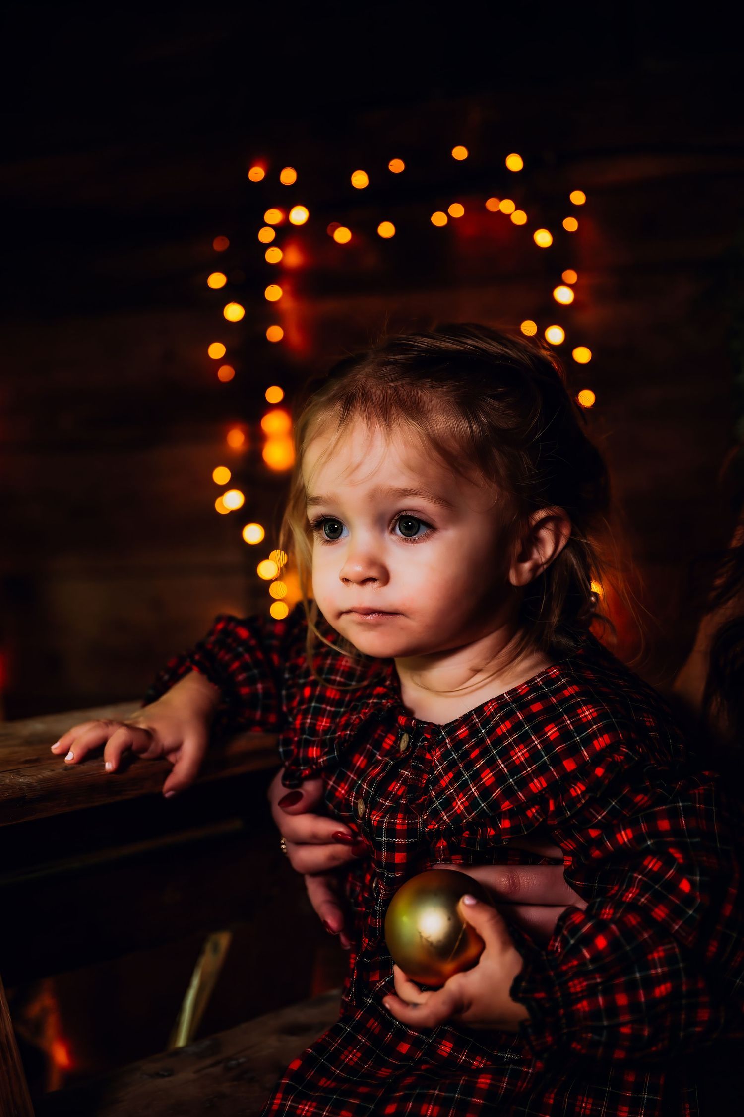Child in red plaid dress holds Christmas ornament while sitting in front of glowing holiday lights.