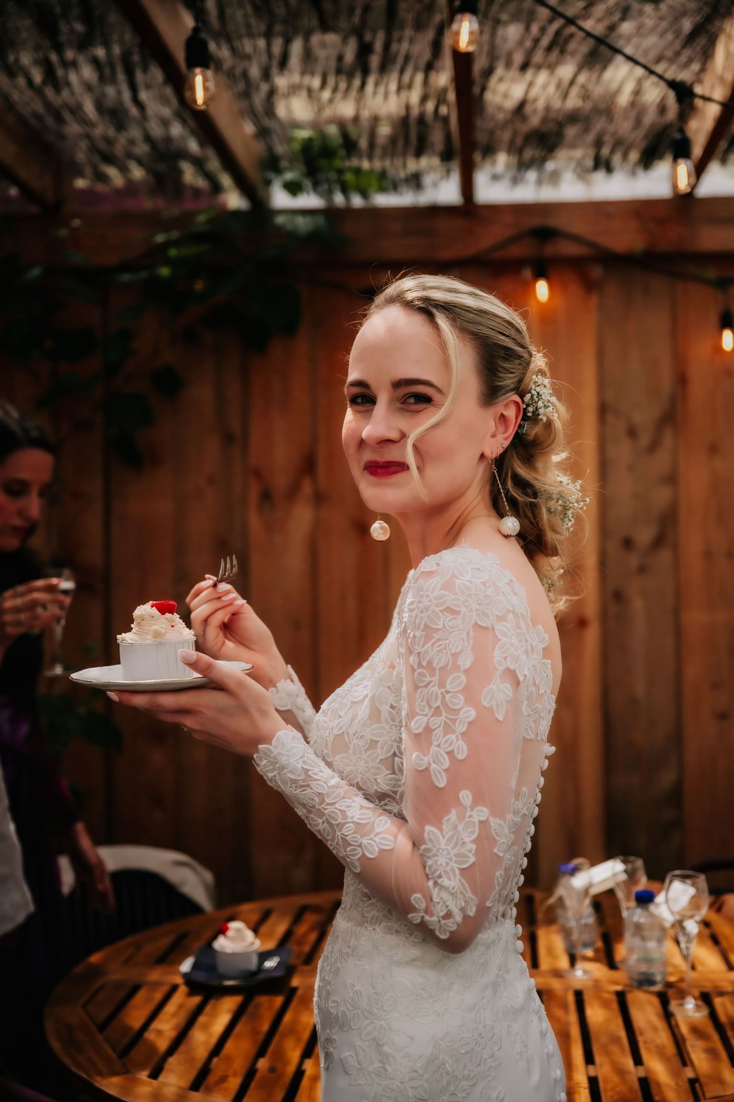 Bride in lace long-sleeve wedding dress holding cake slice on plate, smiling at outdoor rustic wedding reception.