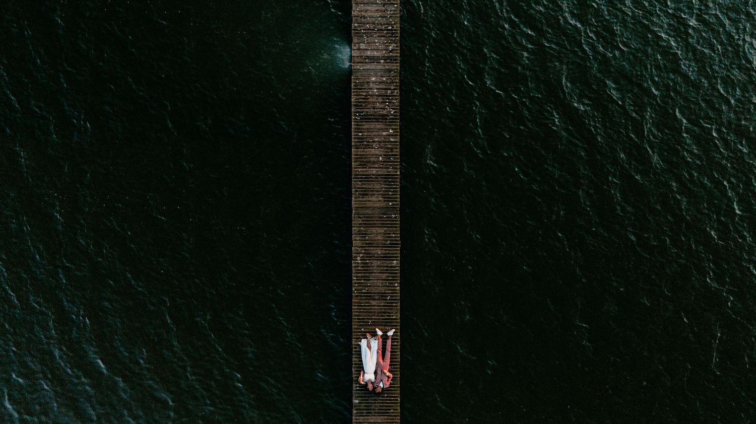 Aerial view of a wooden pier extending into dark teal waters with tiny figures at the end.