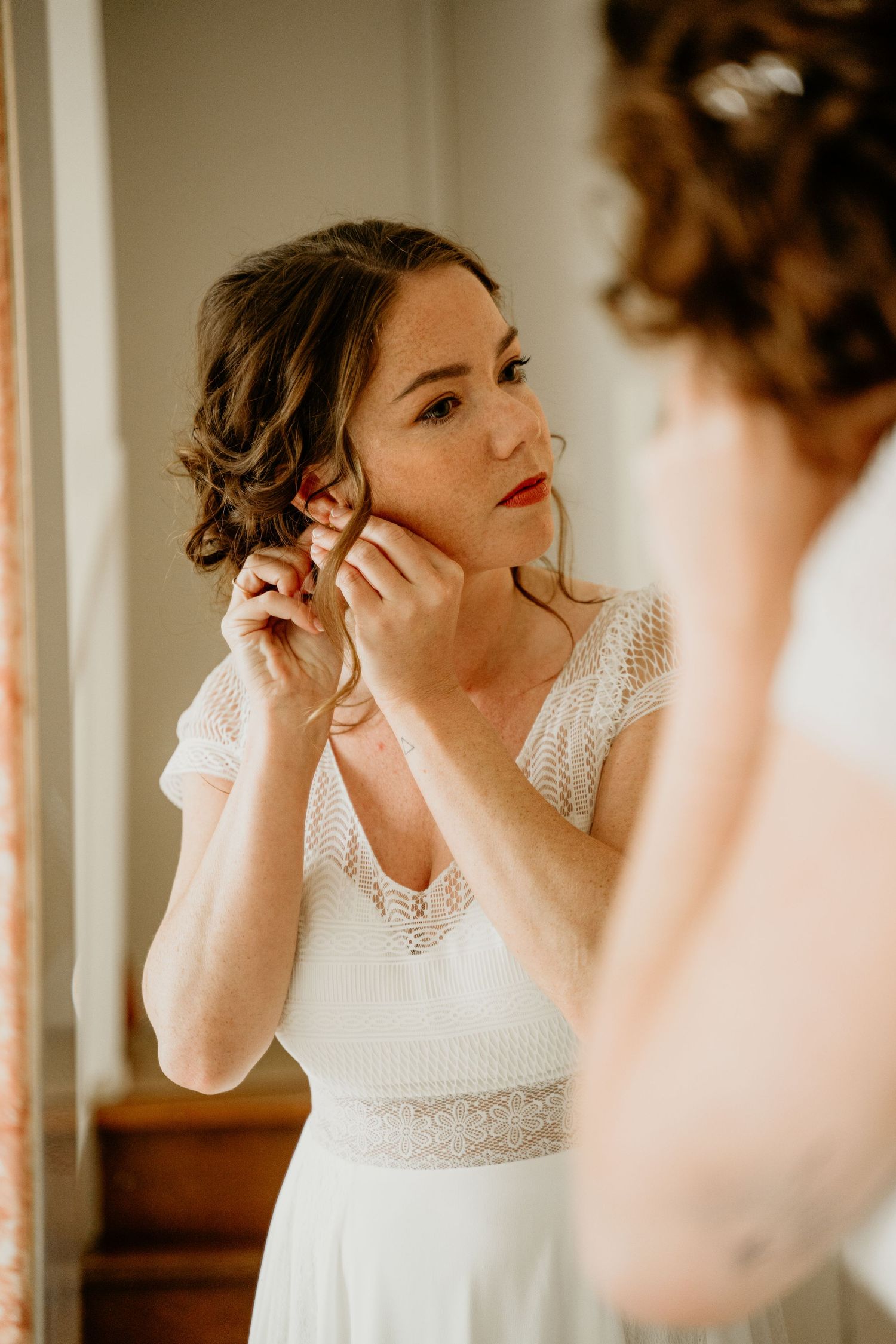 Bride in a white lace dress adjusts her earrings while getting ready in front of a mirror.