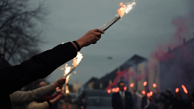 Hands holding burning flares at dusk with glowing red lights in the background during a nighttime gathering.