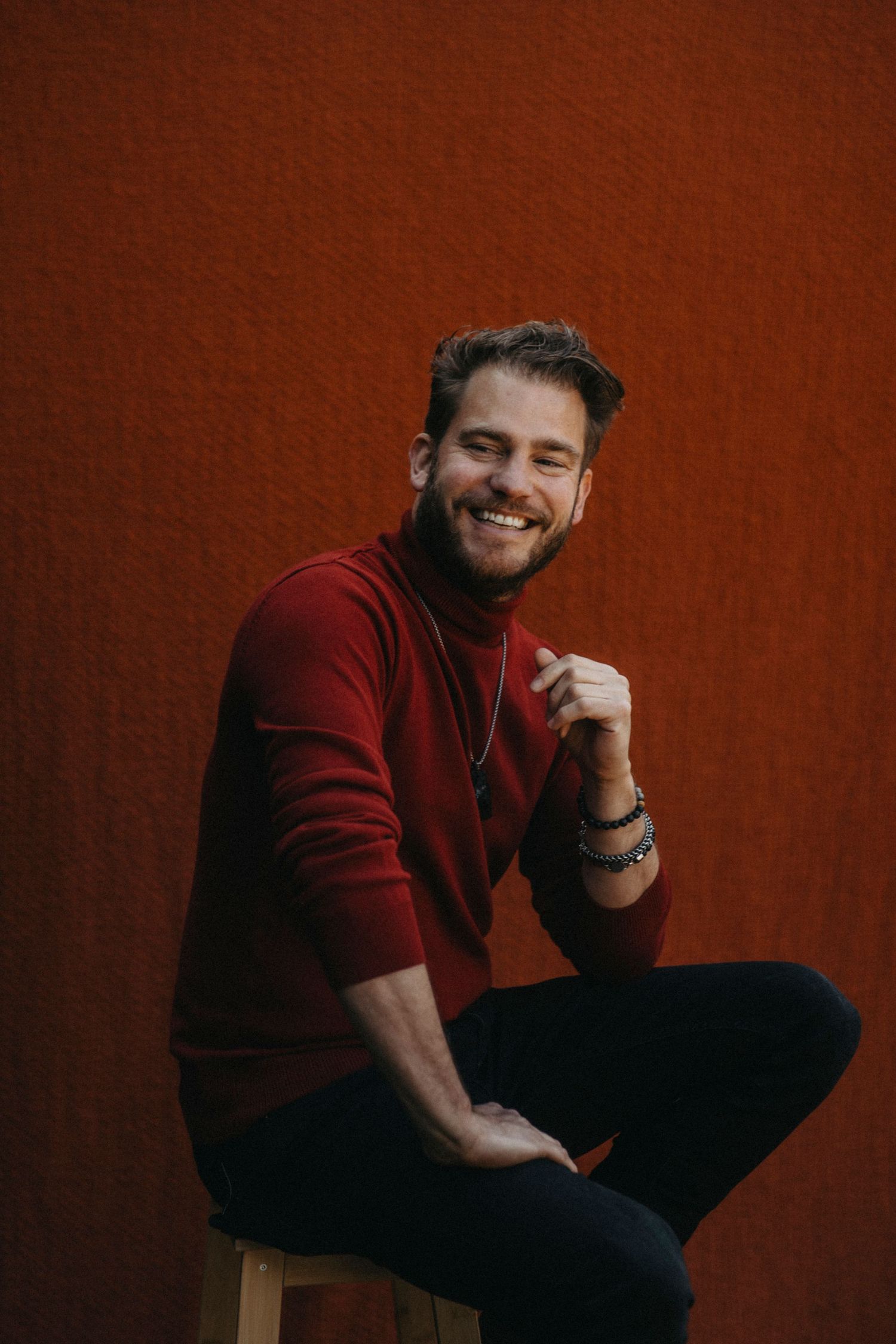 A person in a burgundy sweater sits casually against a dark red background while smiling warmly.