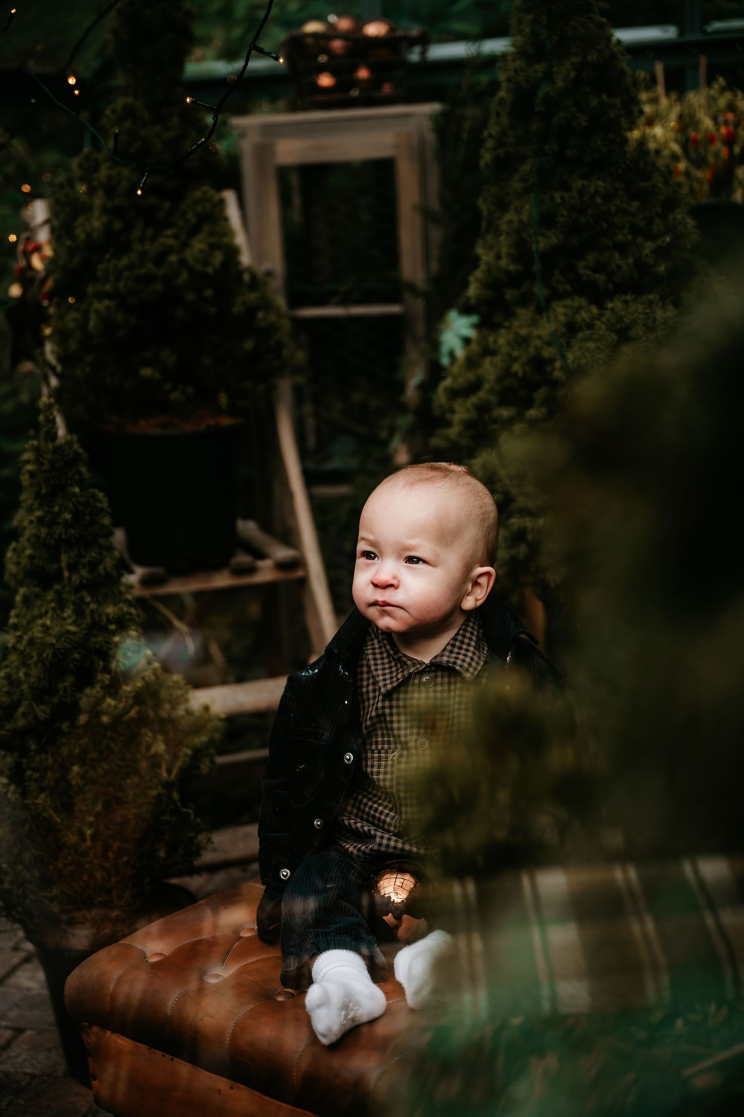 A small child in a dark outfit sits on a brown leather chair in a moody, atmospheric garden setting.