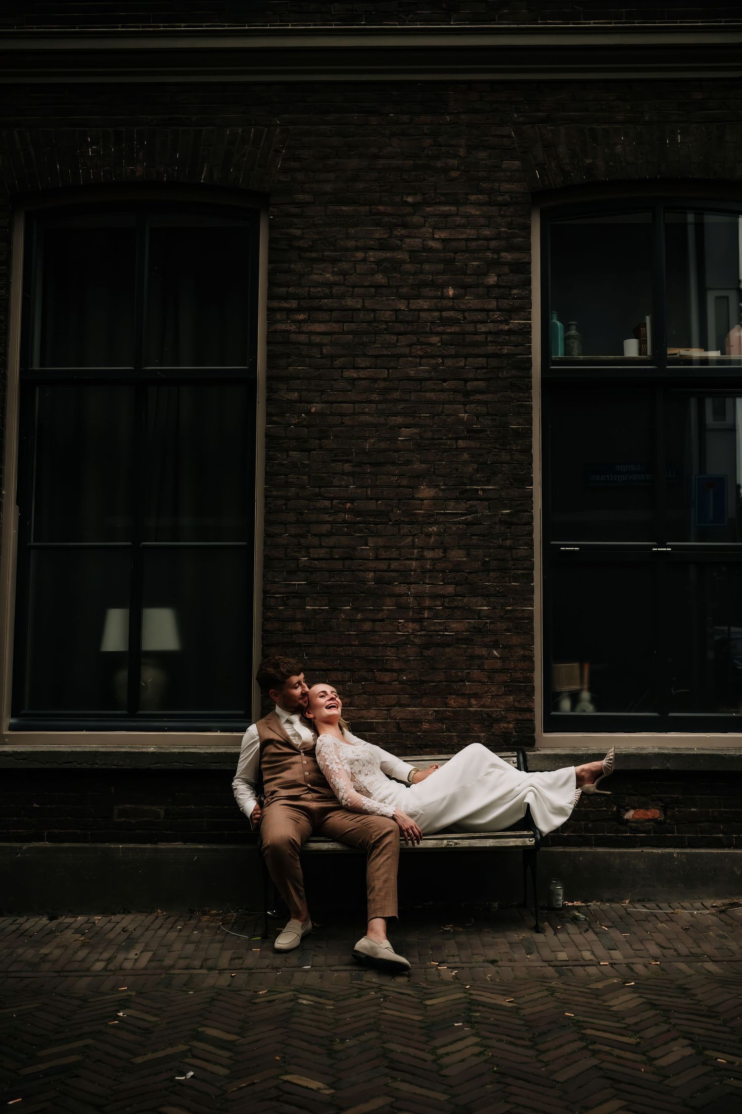 Couple in wedding attire relaxing on a bench against a dark brick wall in a romantic urban setting.