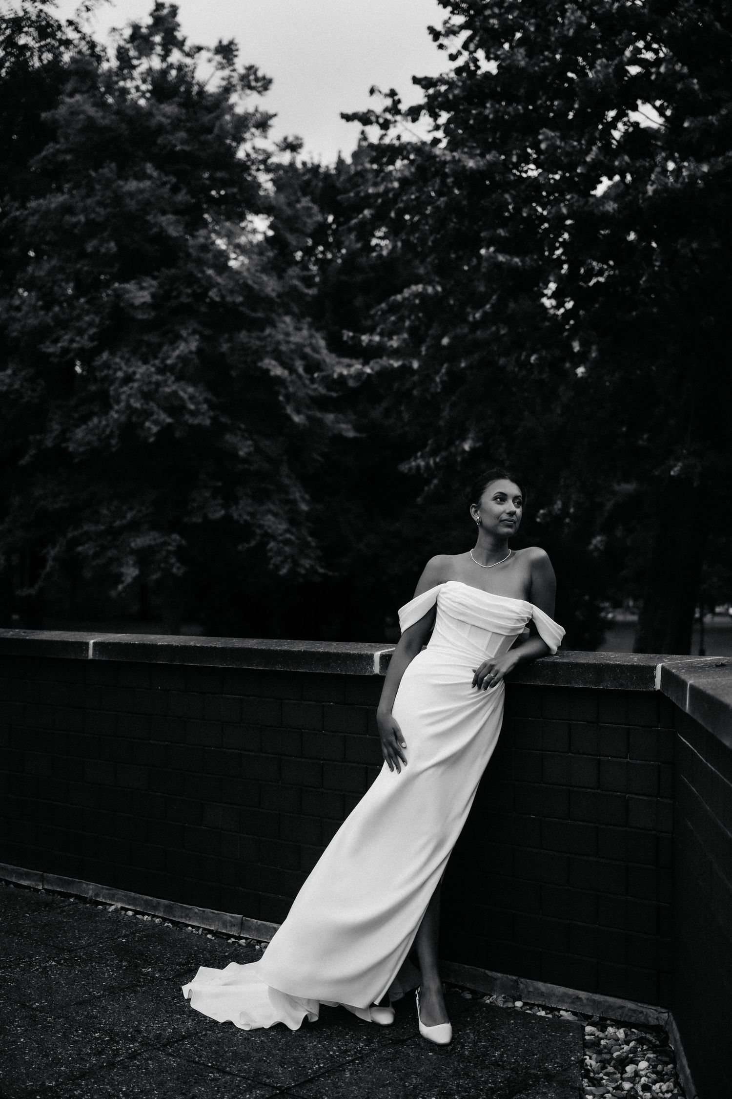 Black and white photo of a bride in an off-shoulder white gown posing gracefully outdoors near a brick wall.