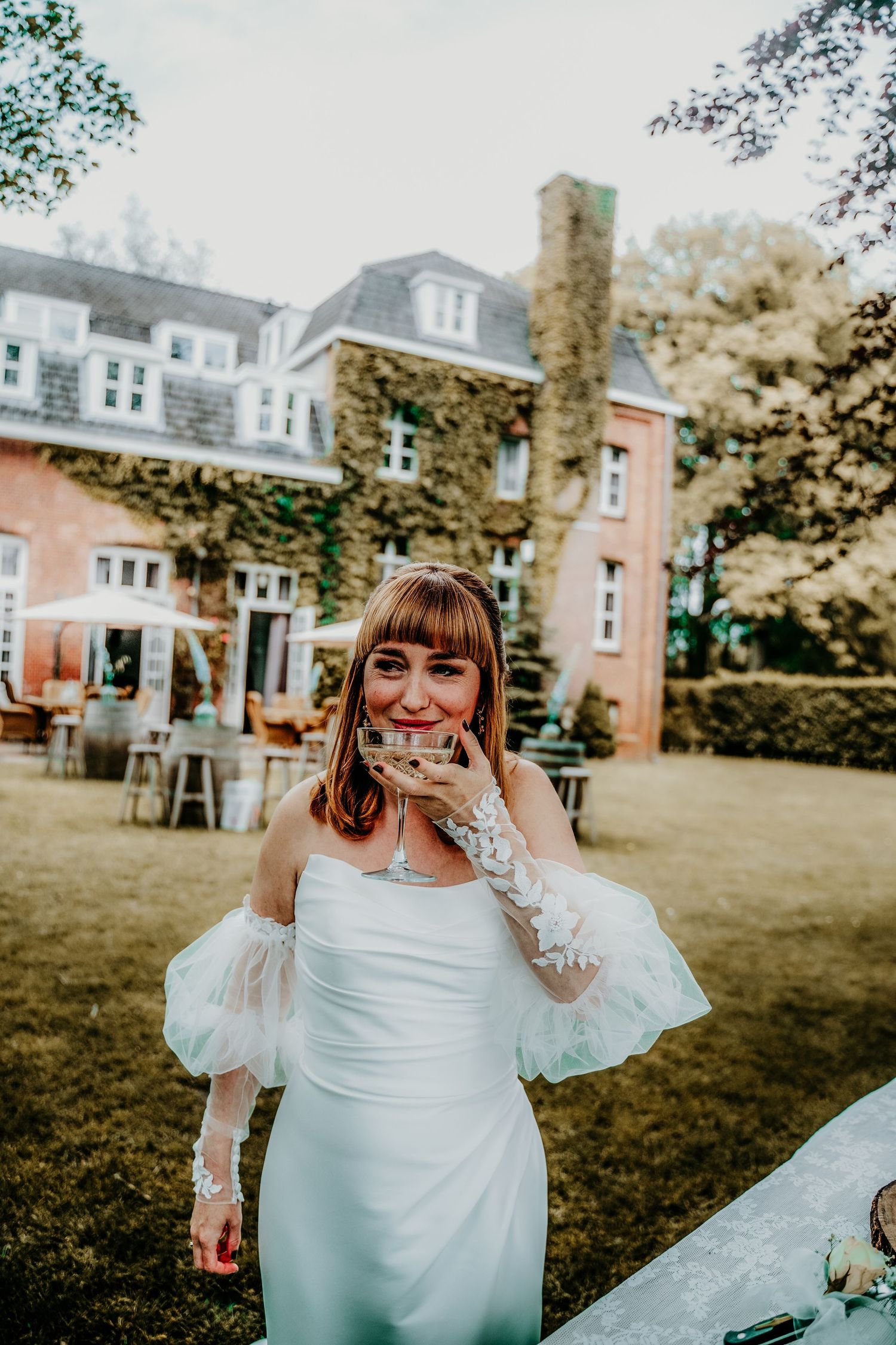 A person in an elegant white wedding gown stands on a lawn in front of a grand historic mansion.