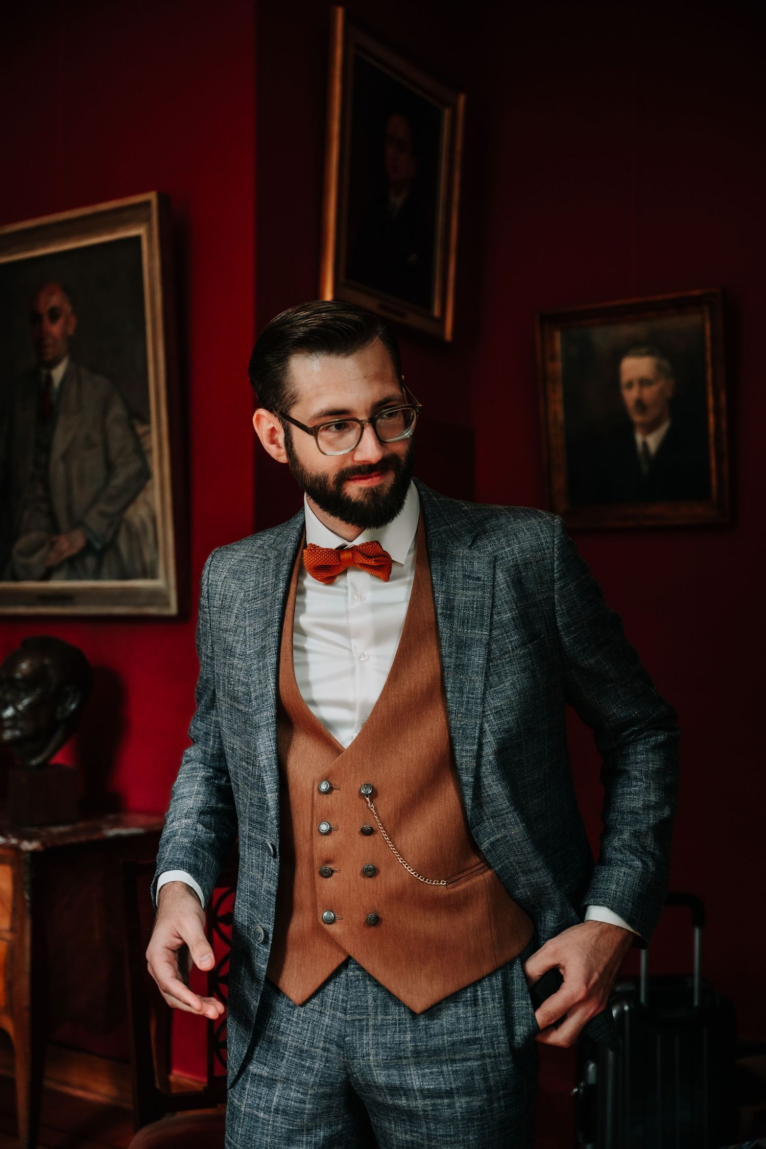 Stylish groom in gray plaid suit and brown vest poses confidently in elegant red room.