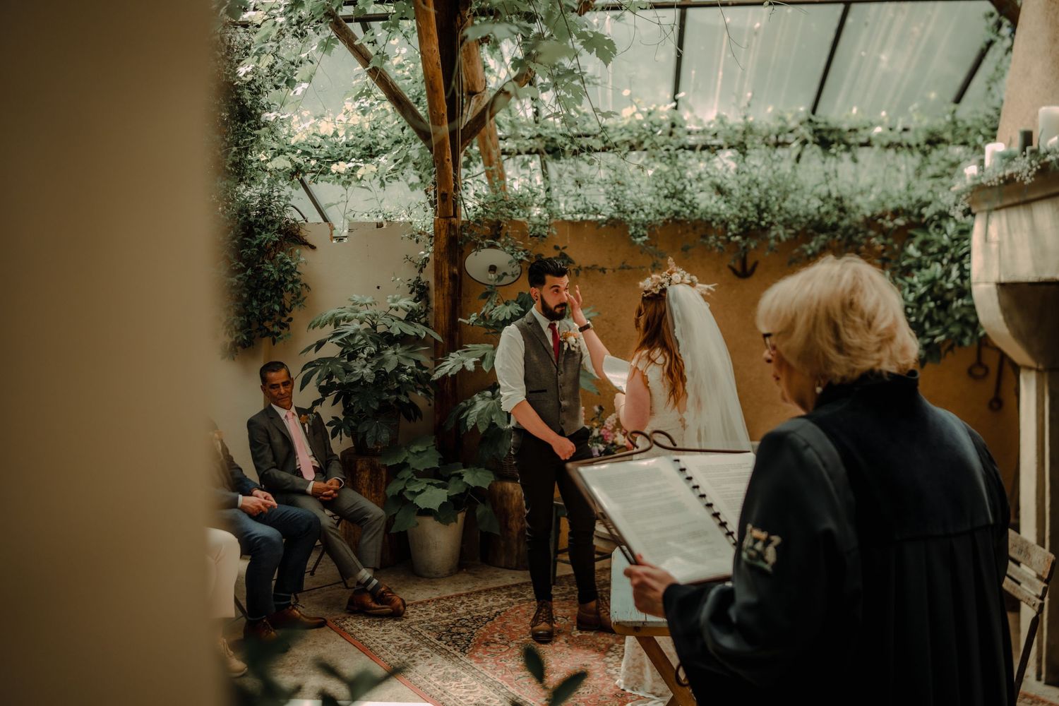 Intimate wedding ceremony in a greenhouse-style venue with natural foliage and rustic decor.