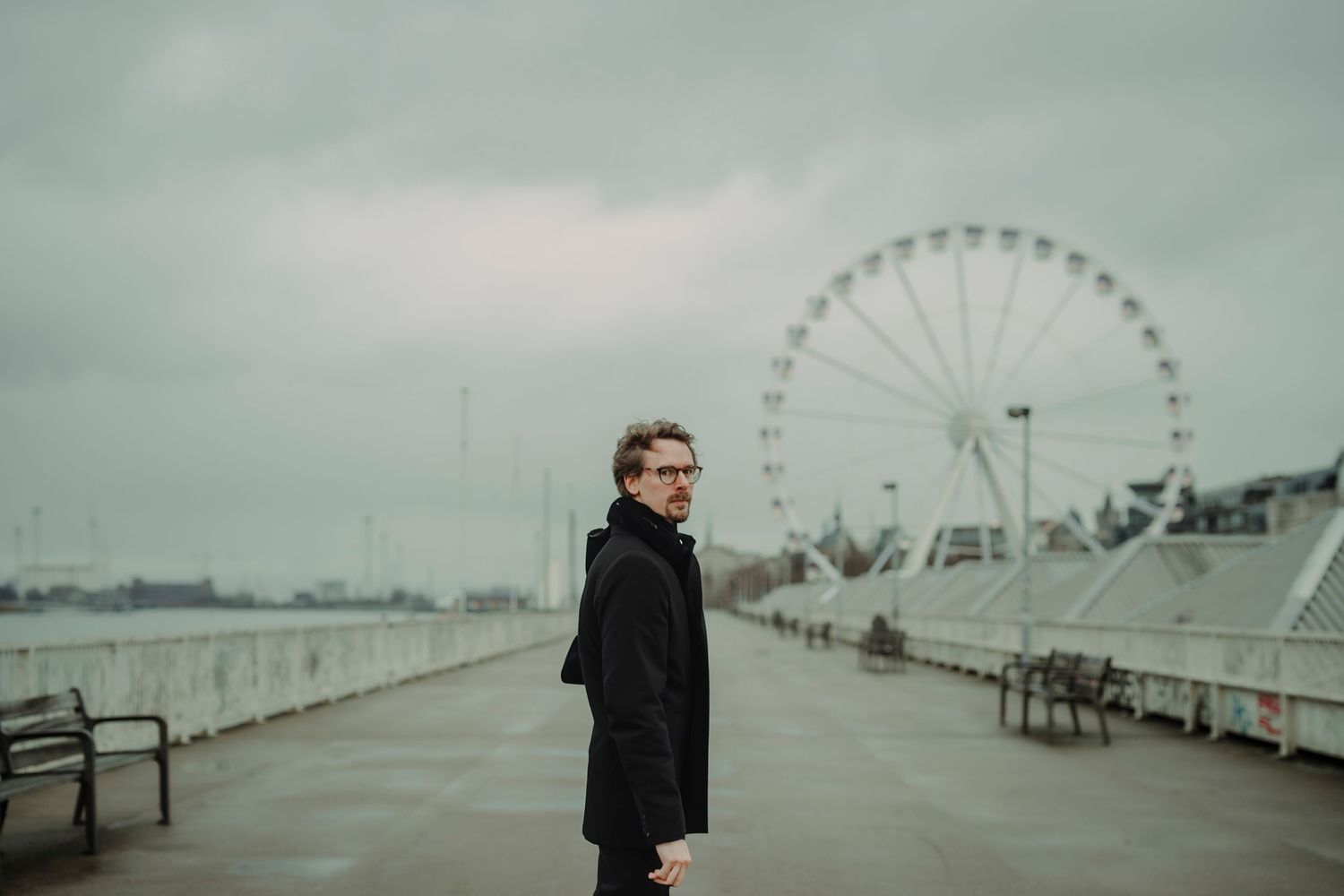 A person in a black coat stands on a rainy waterfront promenade with a large Ferris wheel visible in the background.