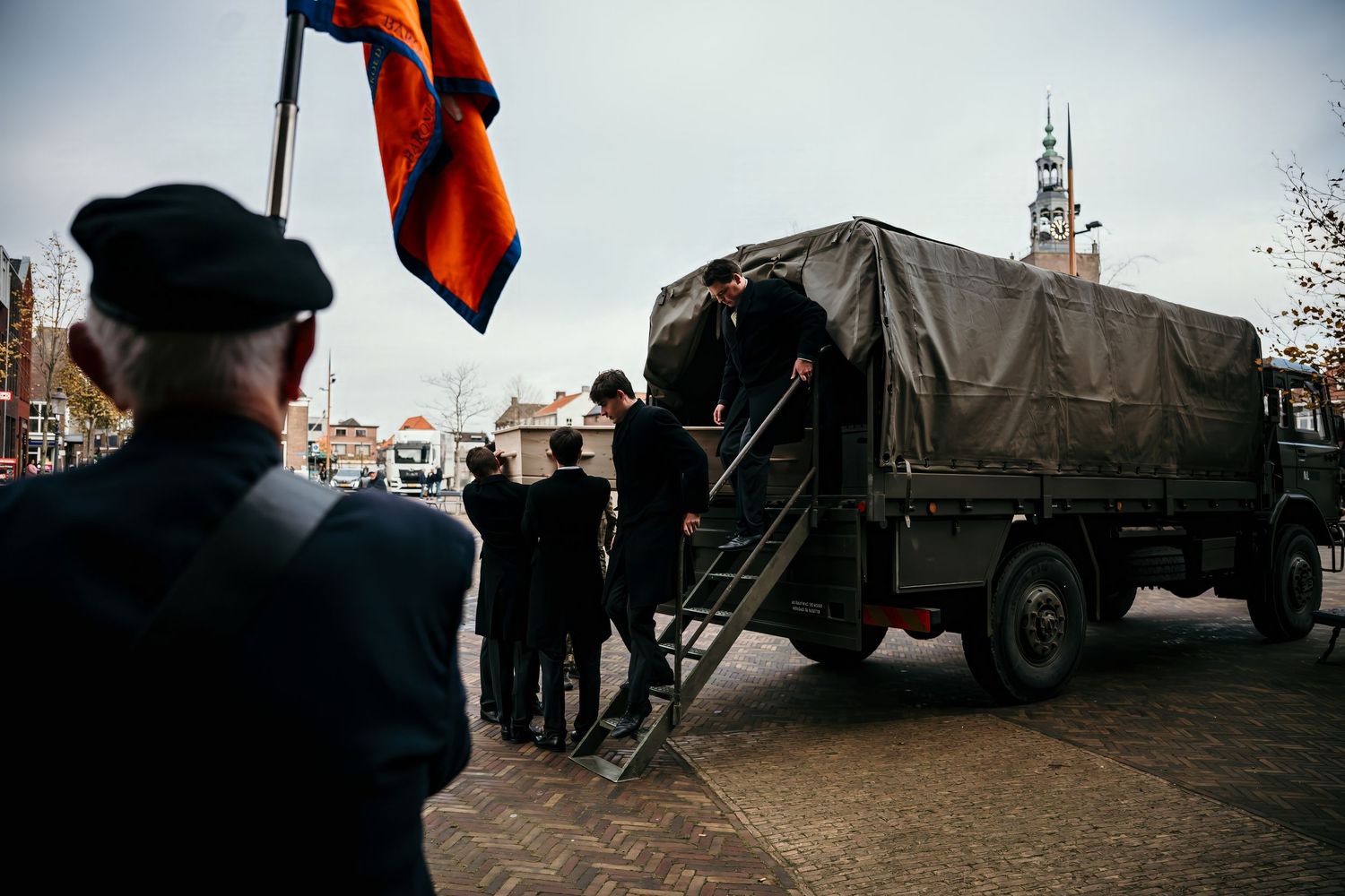 A series of silhouetted figures stand against a bright sky with orange flags and military vehicles in the background.
