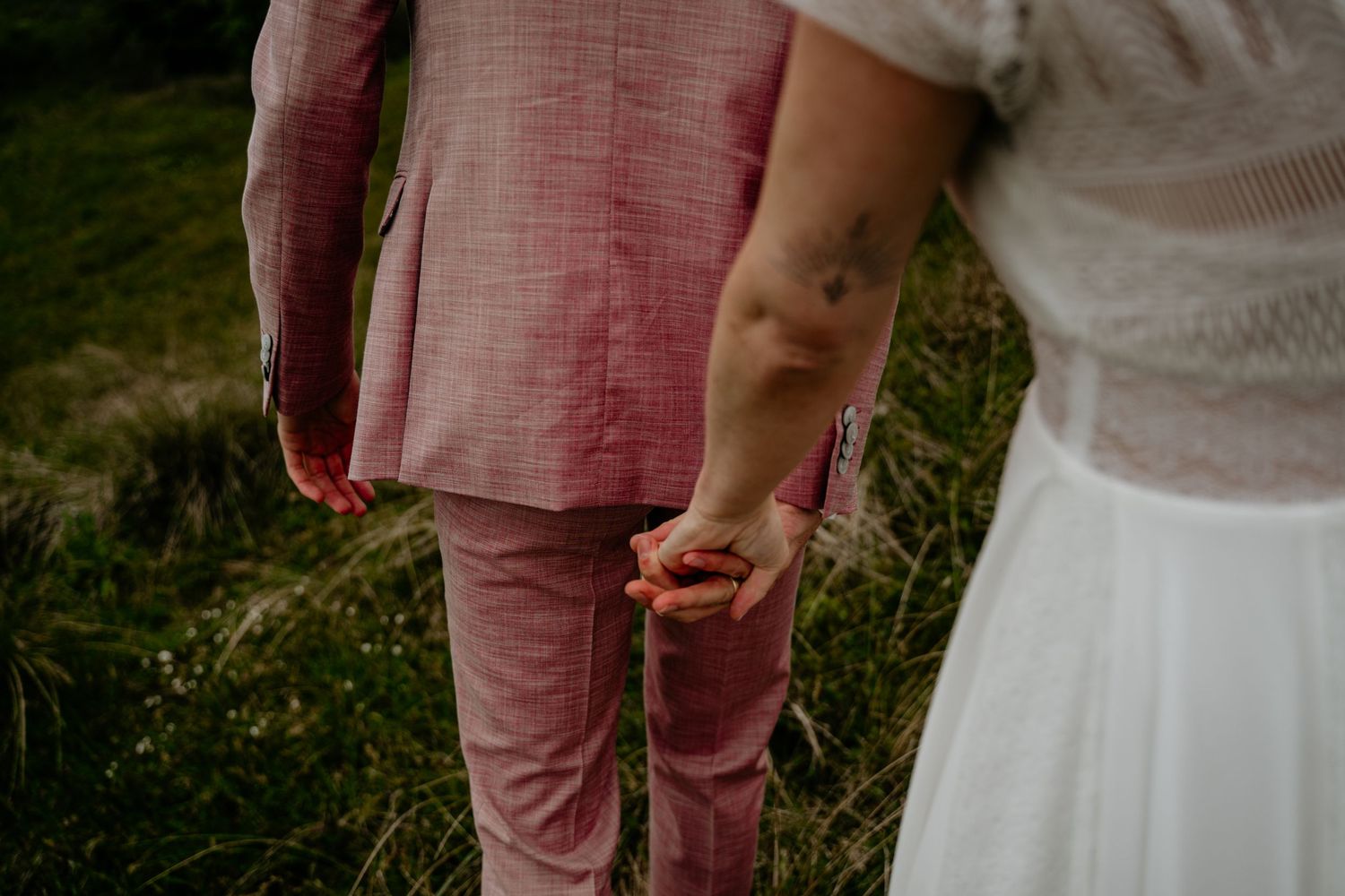 A couple in pink and white outfits hold hands while walking through a grassy outdoor setting.