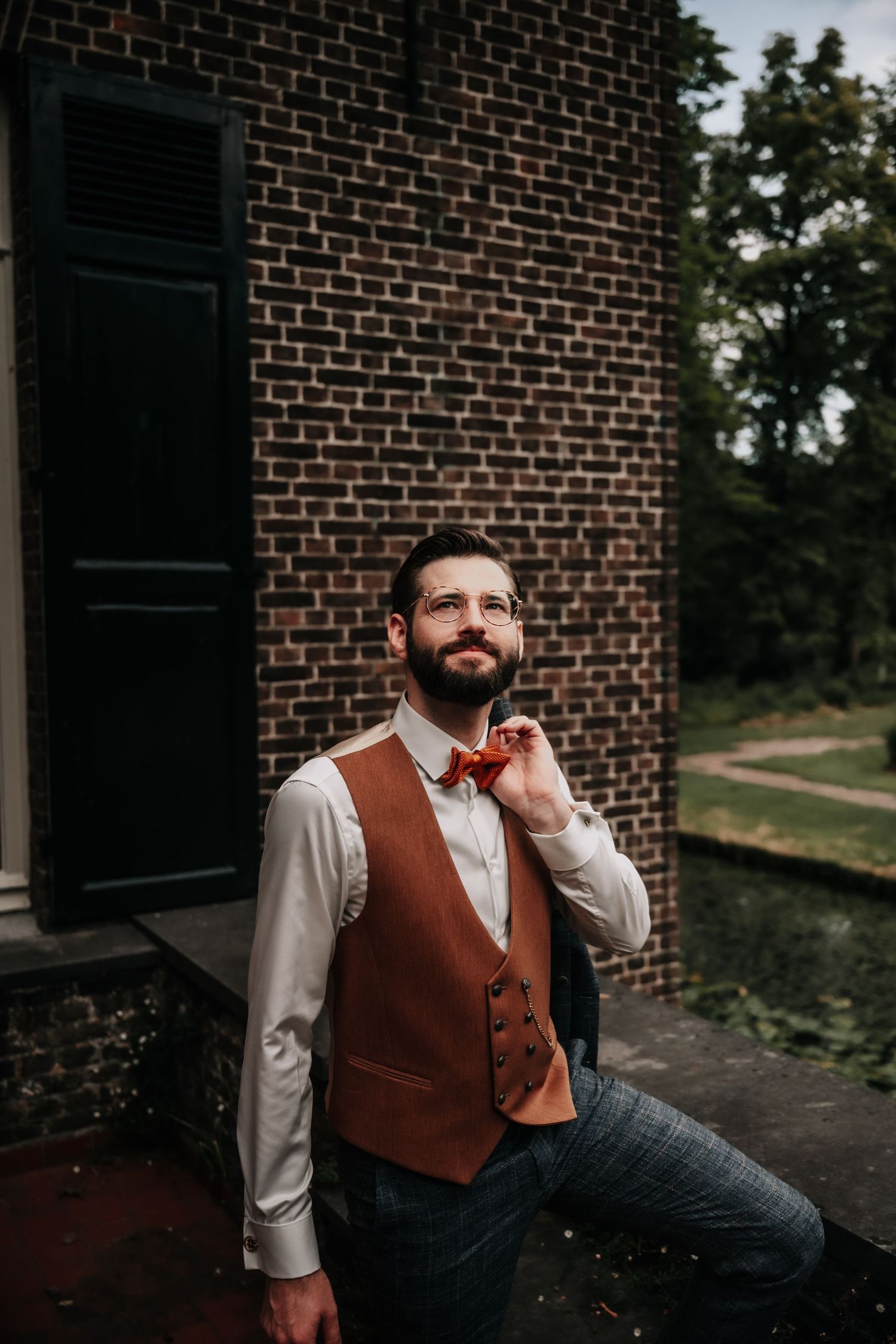 Person in vintage-style vest and dress shirt poses thoughtfully against brick wall exterior.