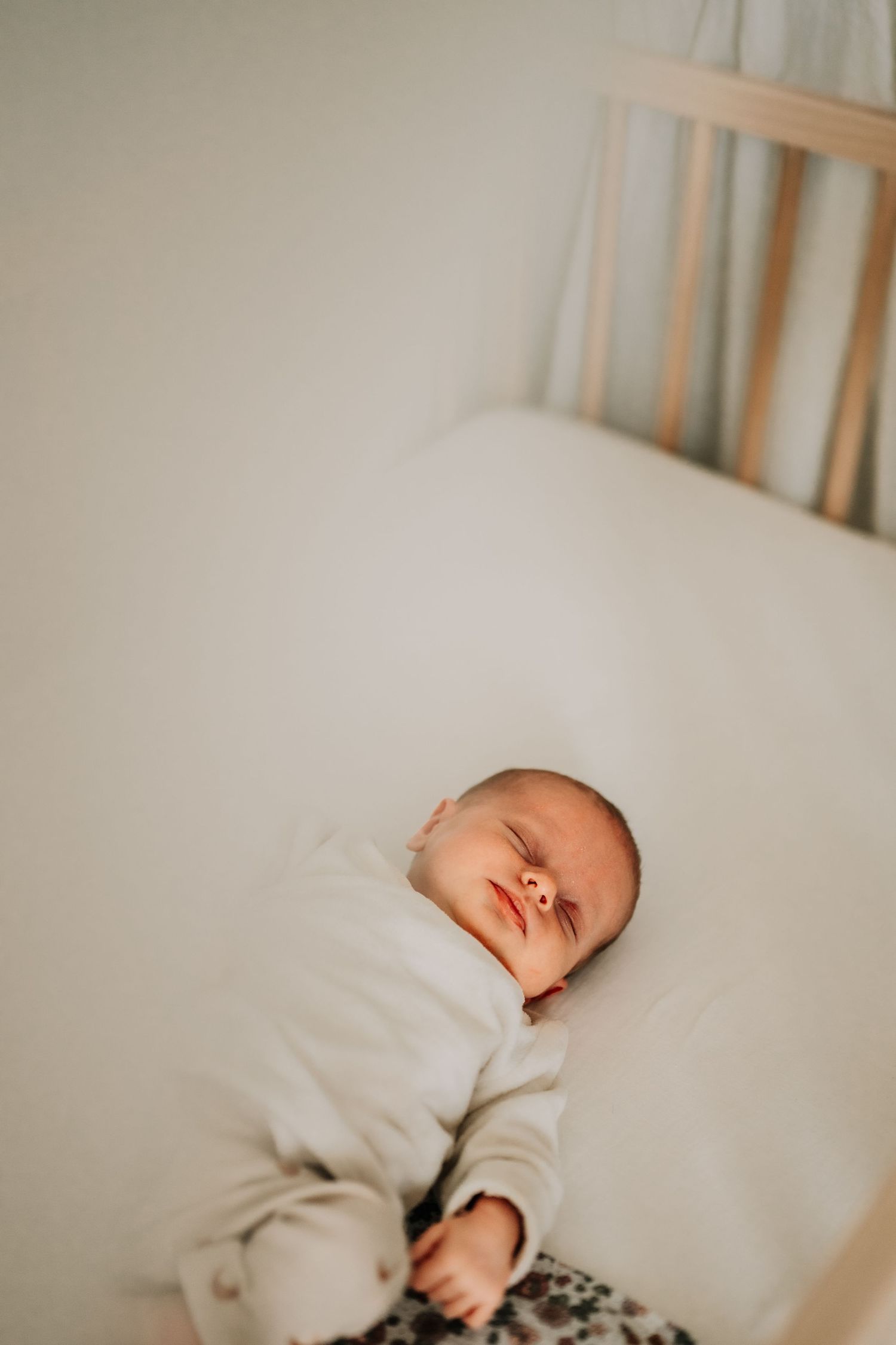 Sleeping baby in white onesie lying peacefully in a wooden crib with soft bedding.