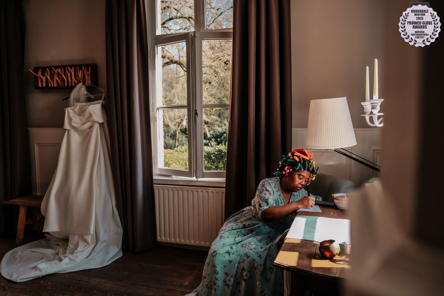 A wedding dress hangs by a window while someone in a teal dress writes at an antique desk in a cozy room.