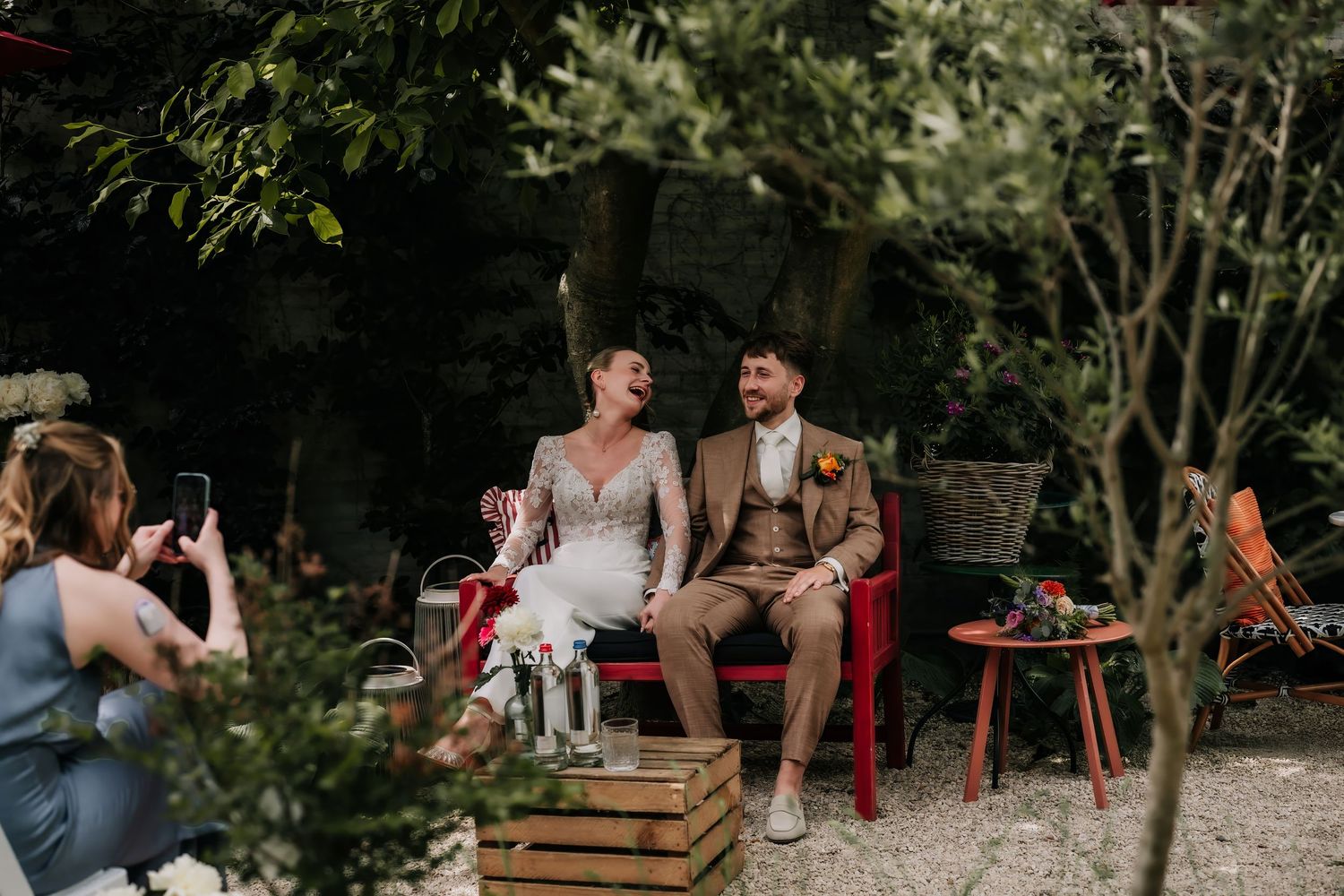 Bride and groom sitting on a red bench laughing during outdoor wedding ceremony while guest photographs them.