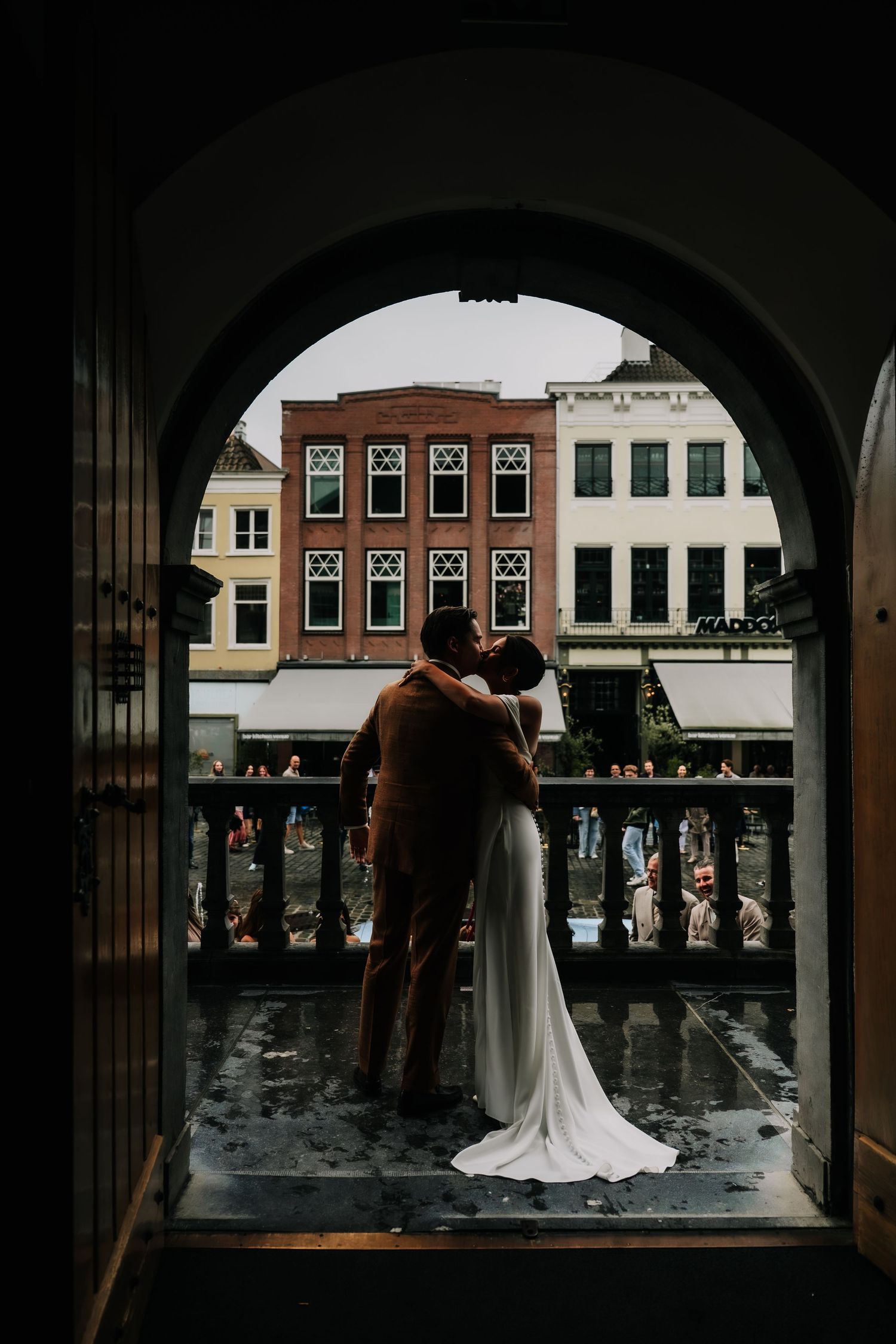 Couple sharing a kiss in an arched doorway, bride in white gown, groom in brown suit, with European buildings in background.