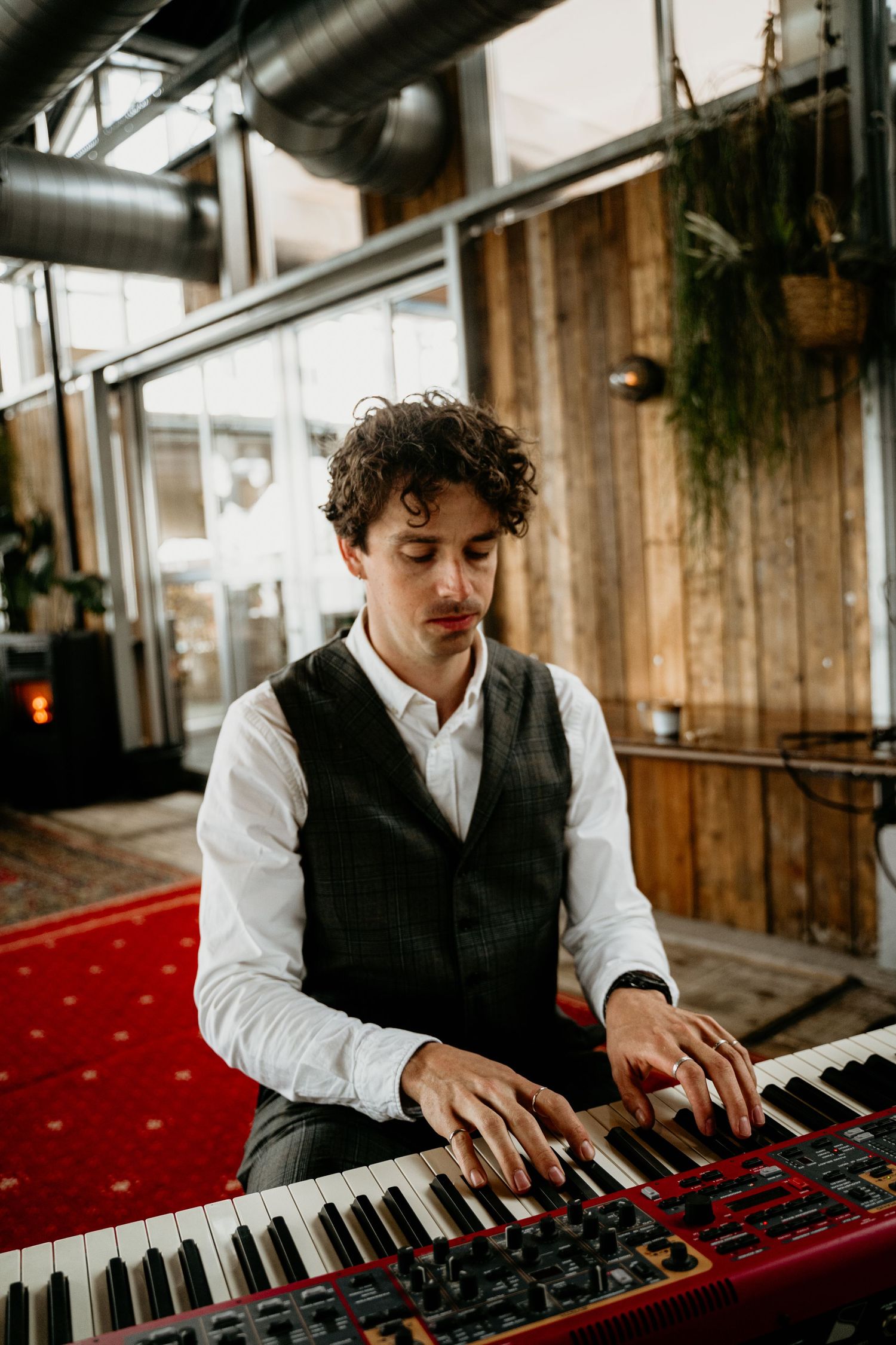 Musician in a black vest and white shirt plays a red piano keyboard in an industrial-style venue.