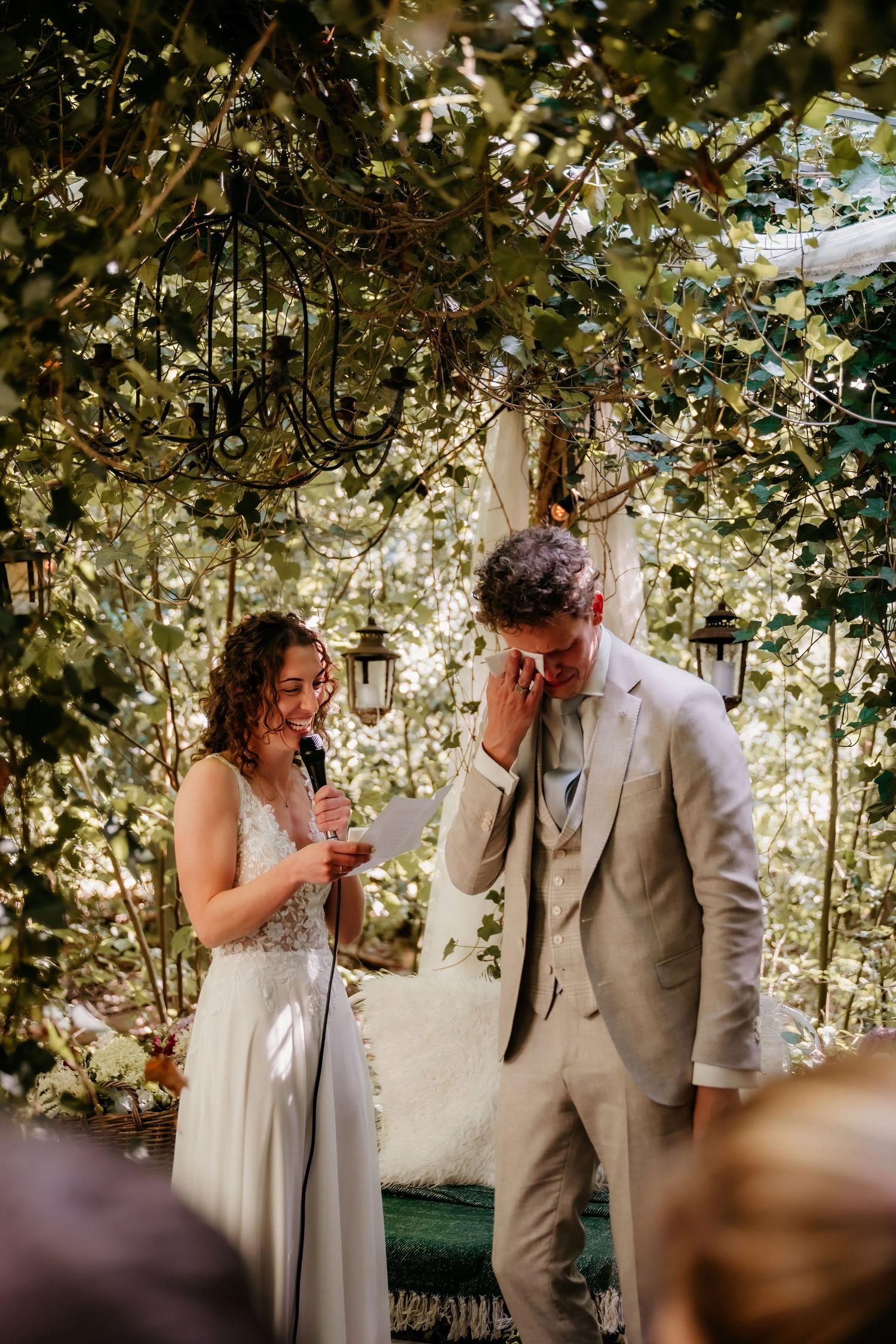 A couple shares a joyful moment under a canopy of white flowers and greenery during an outdoor wedding ceremony.