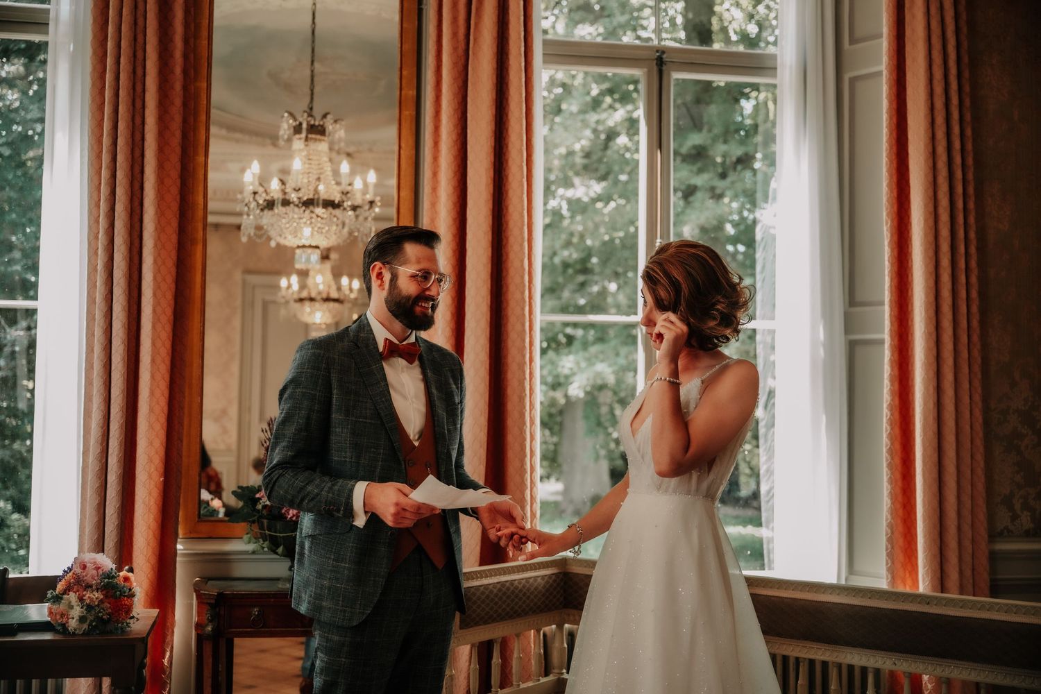 A wedding couple shares an intimate moment in an elegant room with large windows and chandeliers.