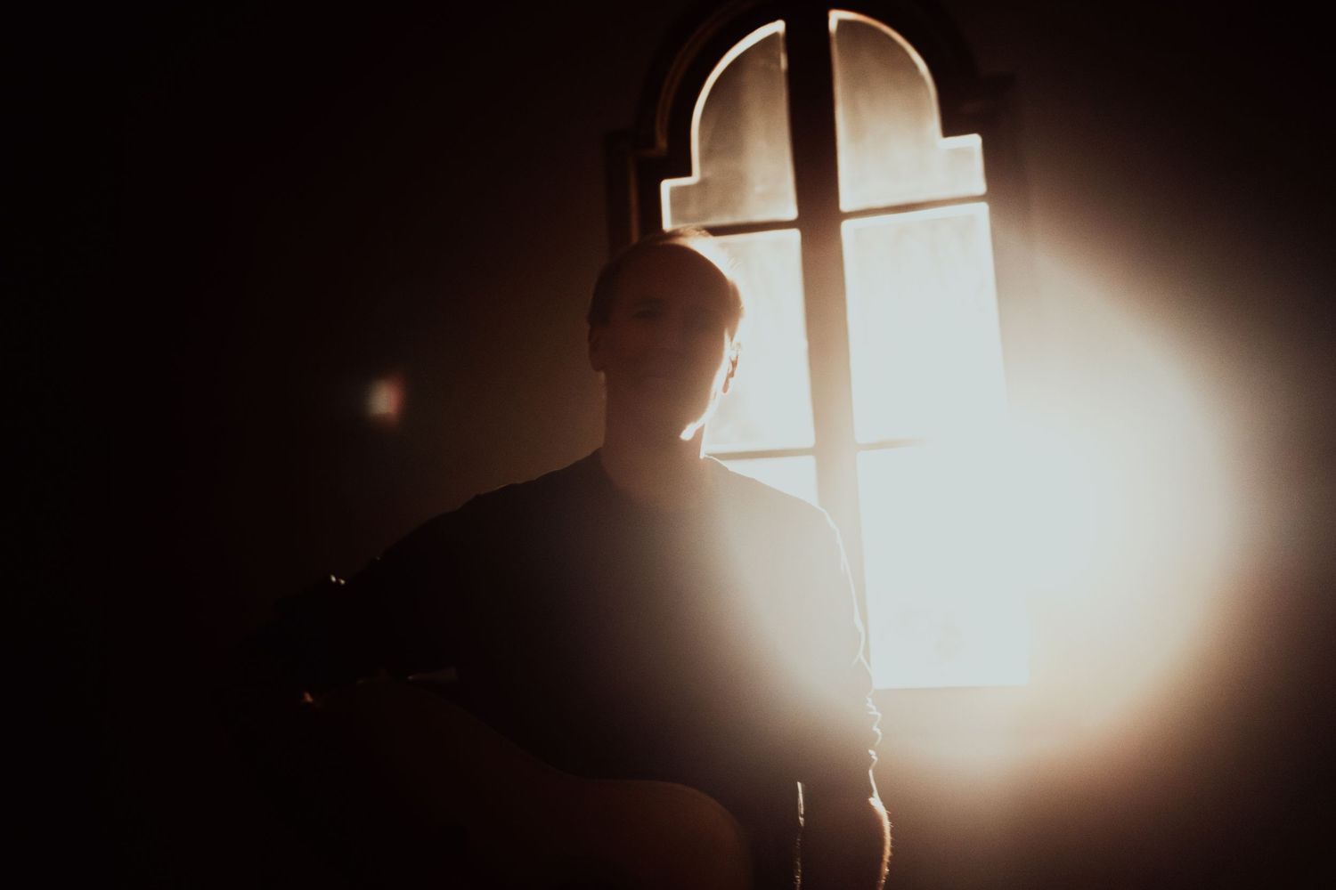 A silhouetted figure stands by a window with bright sunlight streaming through, creating a dramatic backlit effect.