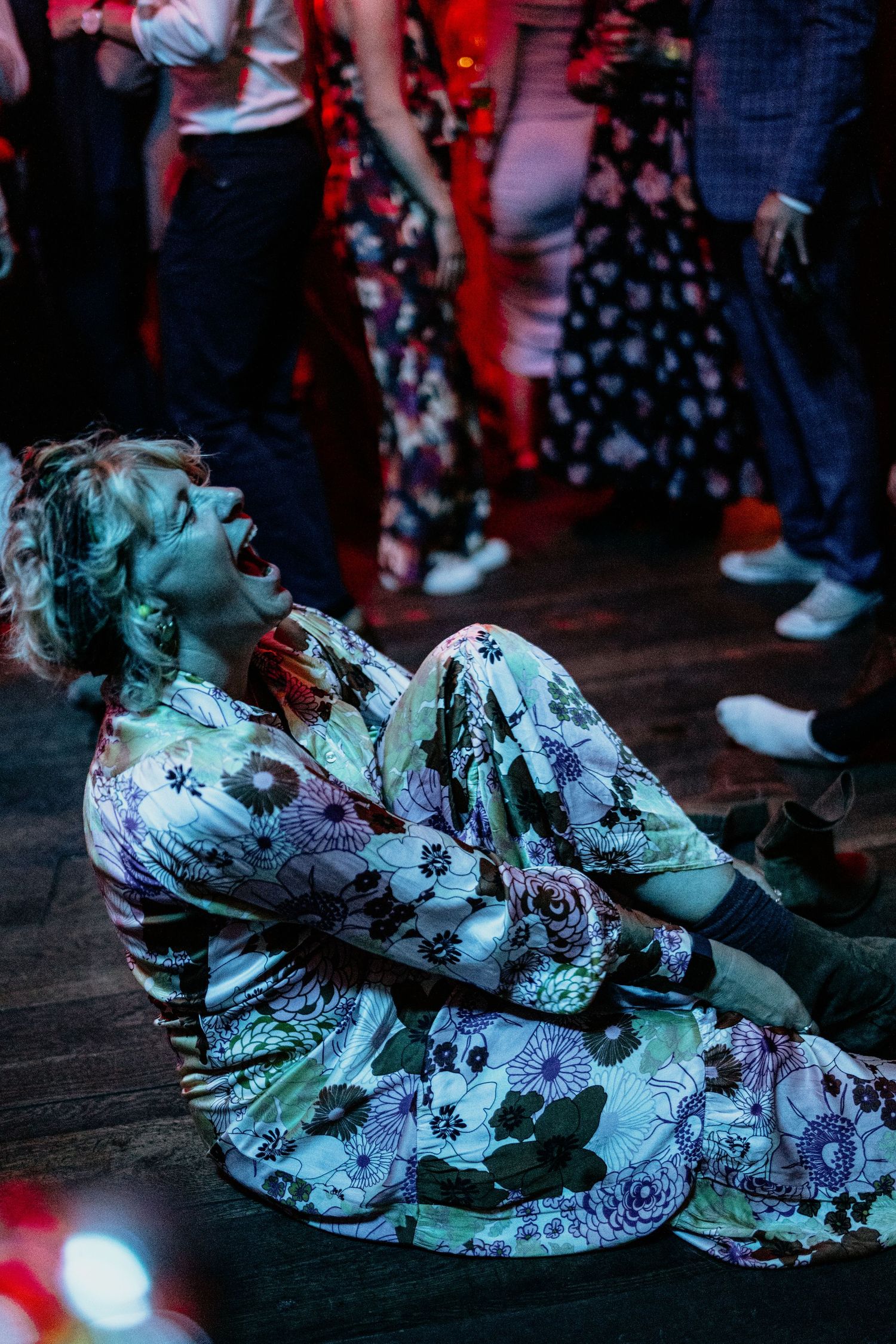 Someone in a floral outfit lies on a dance floor during a party under red lighting.