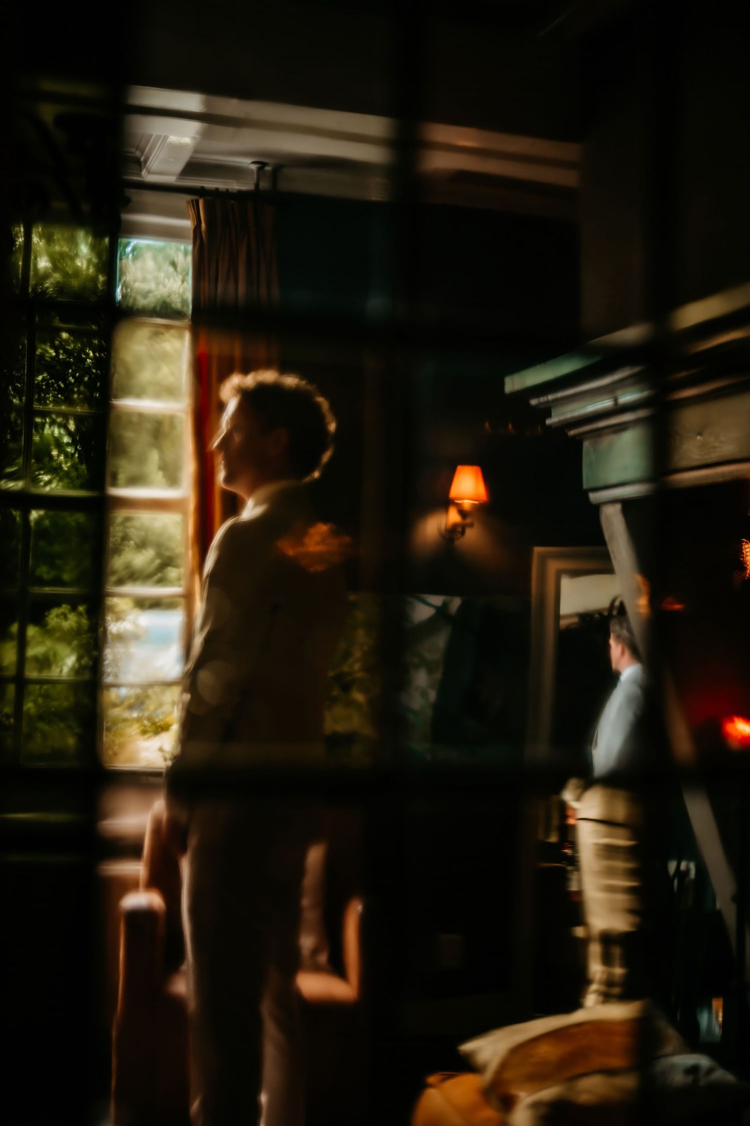 Silhouette figure stands near window in moody interior with warm lamp light creating atmospheric shadows.