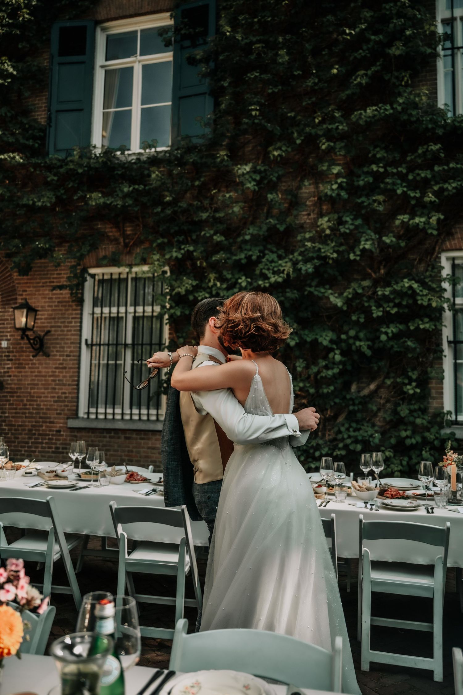 An outdoor wedding reception setup with white chairs and ivy-covered brick walls.