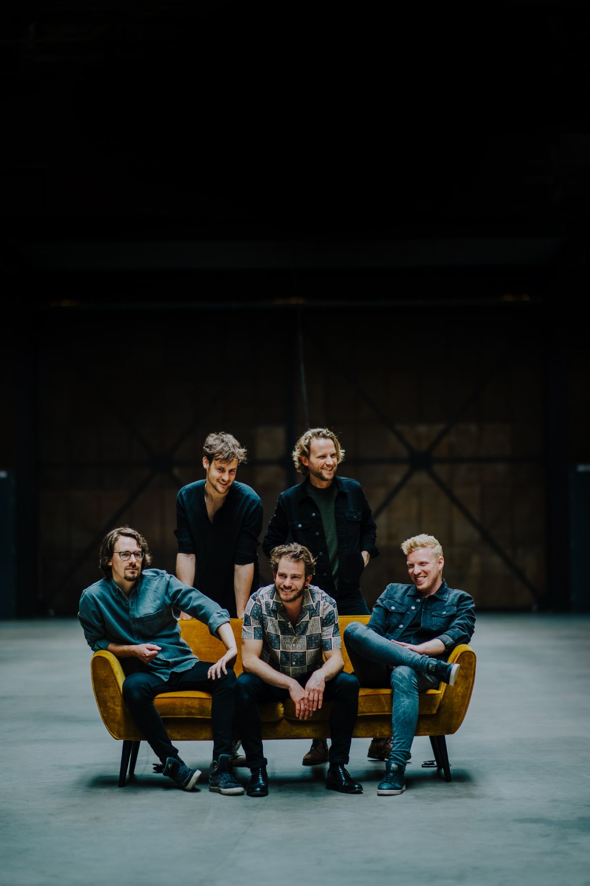 Four people sit casually on a yellow couch in a dark studio space, wearing casual clothing and sneakers.