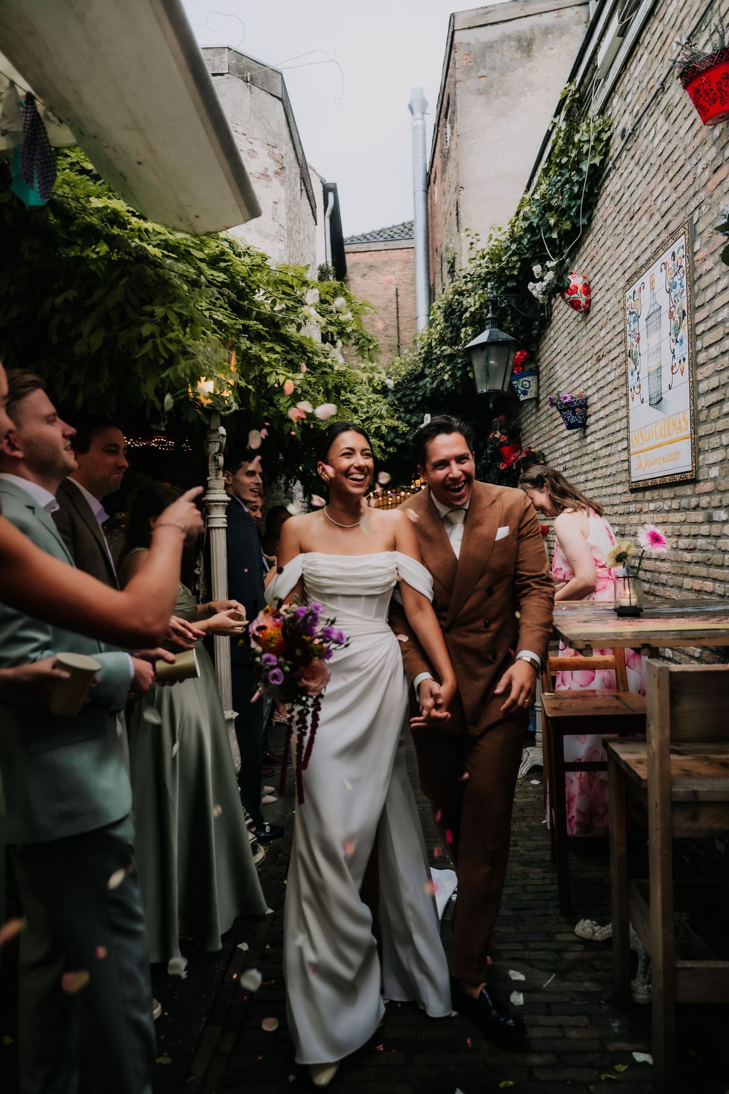 Joyful newlyweds walk through a charming outdoor alley as guests toss confetti, bride holding colorful bouquet.