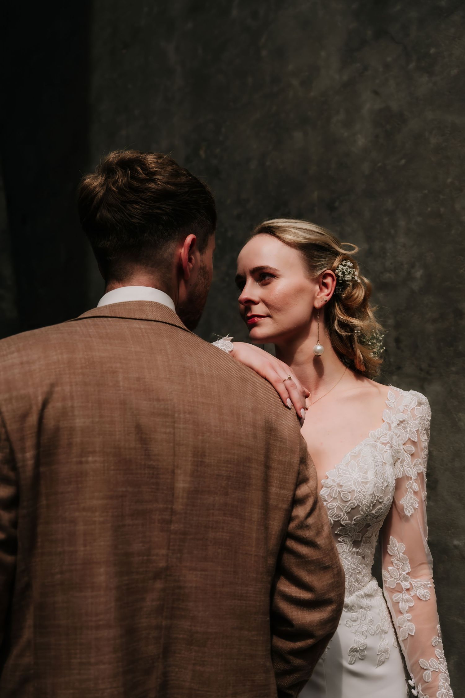 Bride in lace long-sleeve gown gazes at groom in brown suit during intimate wedding portrait against dark backdrop.