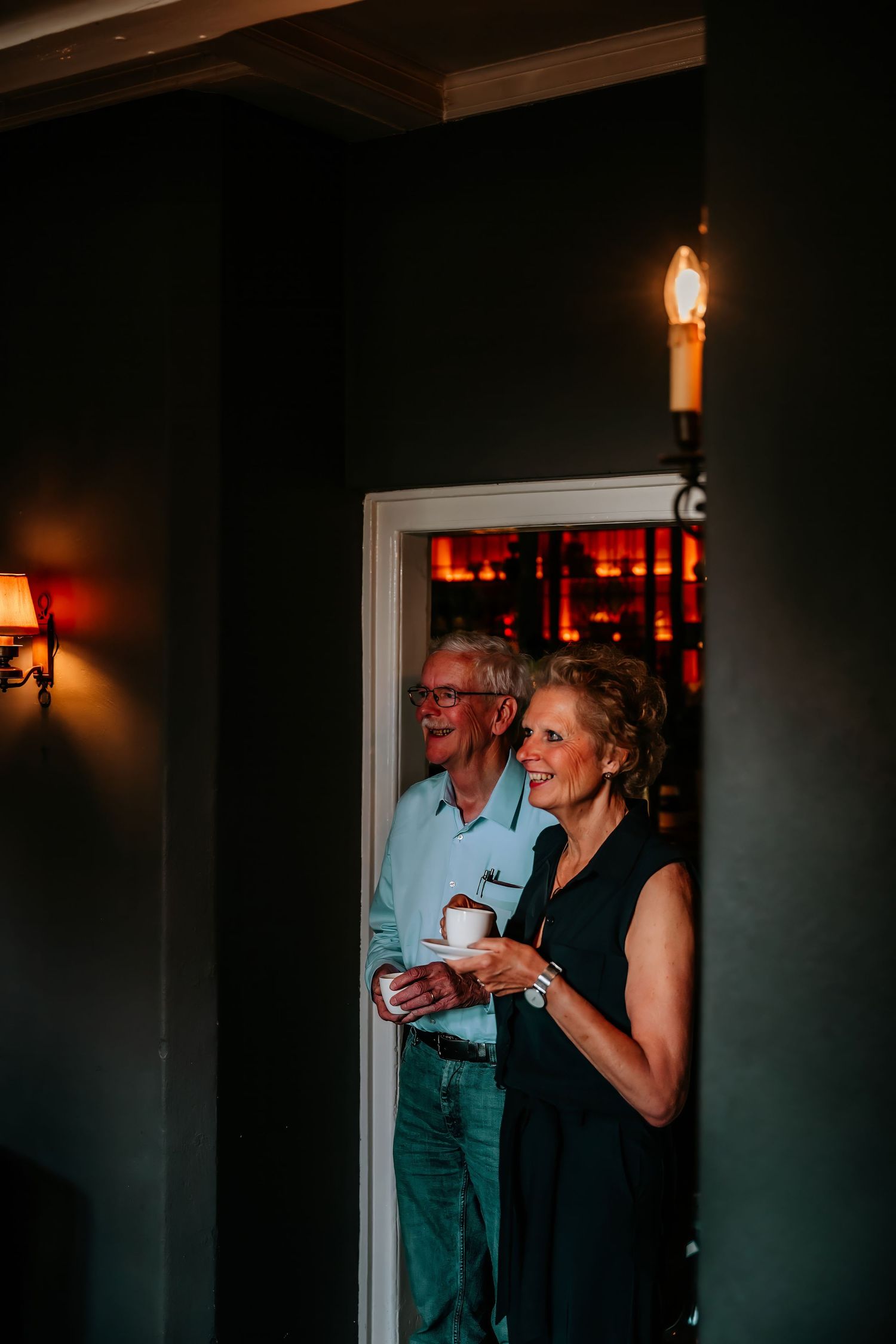 Two guests share a laugh in a dimly lit hallway with warm lighting and dark walls.