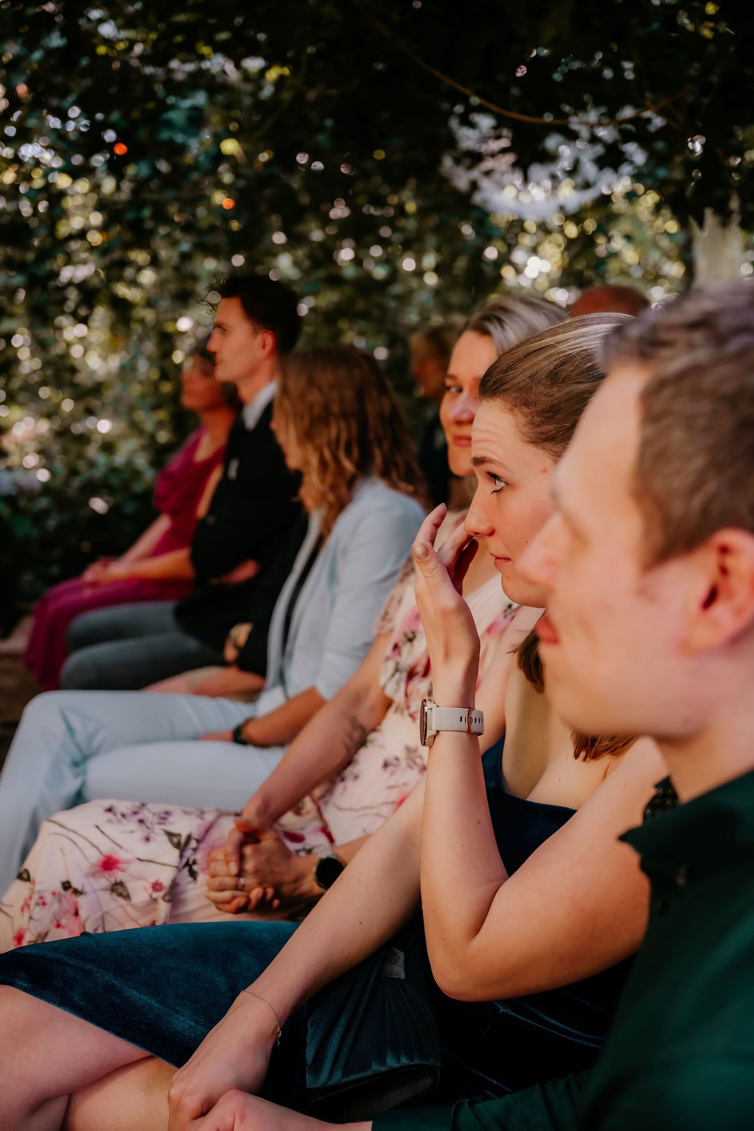 Emotional guests react during an outdoor ceremony while seated in rows beneath trees.