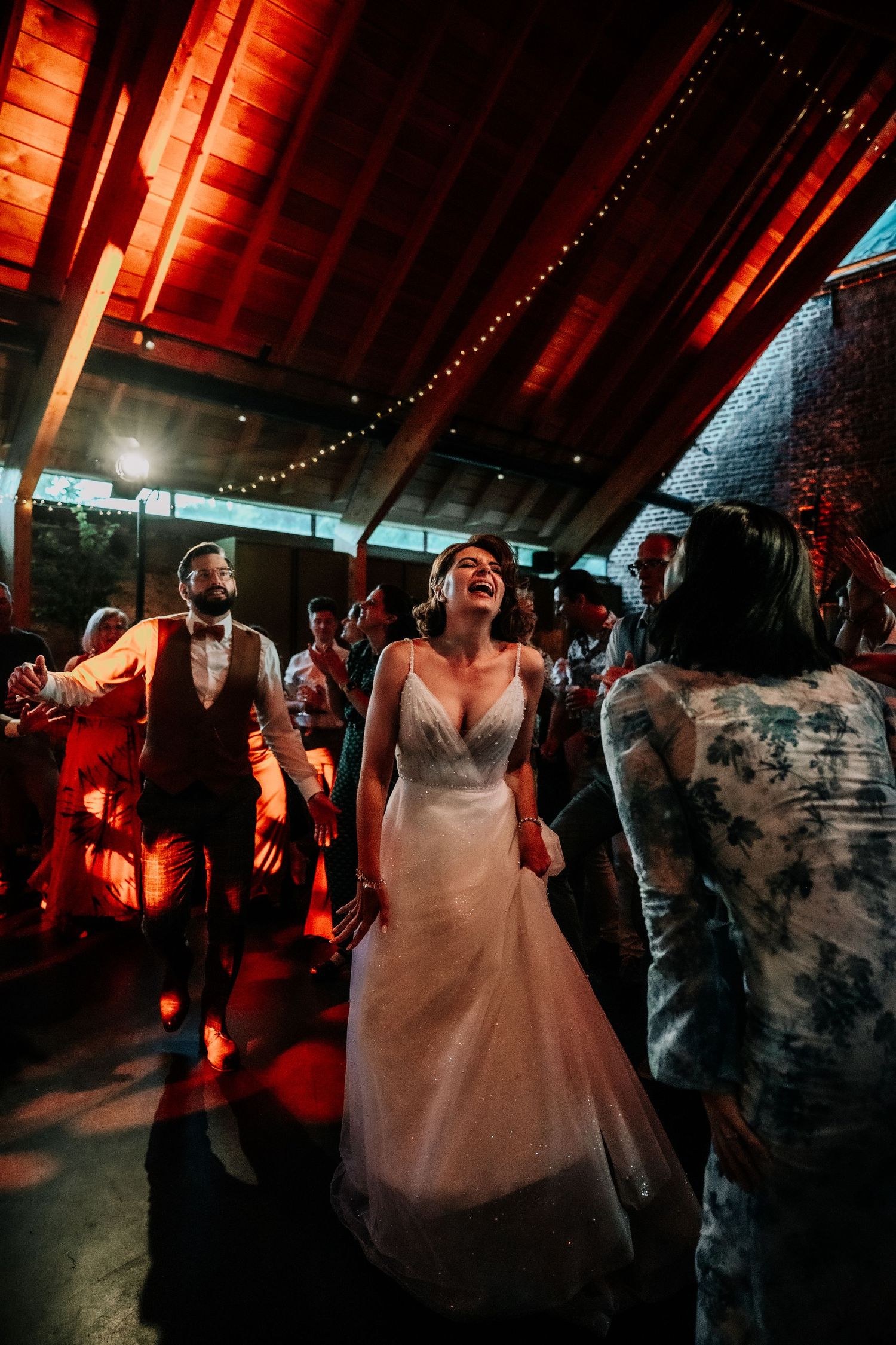 Wedding reception dance party in a rustic barn venue with red ambient lighting and string lights.