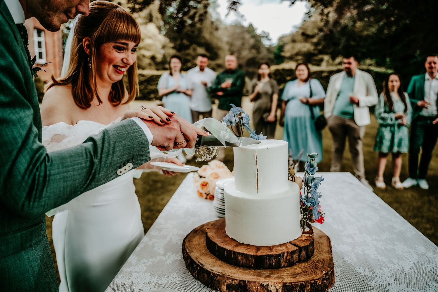 Rustic wedding cake cutting ceremony on wooden slice stand with natural outdoor backdrop.