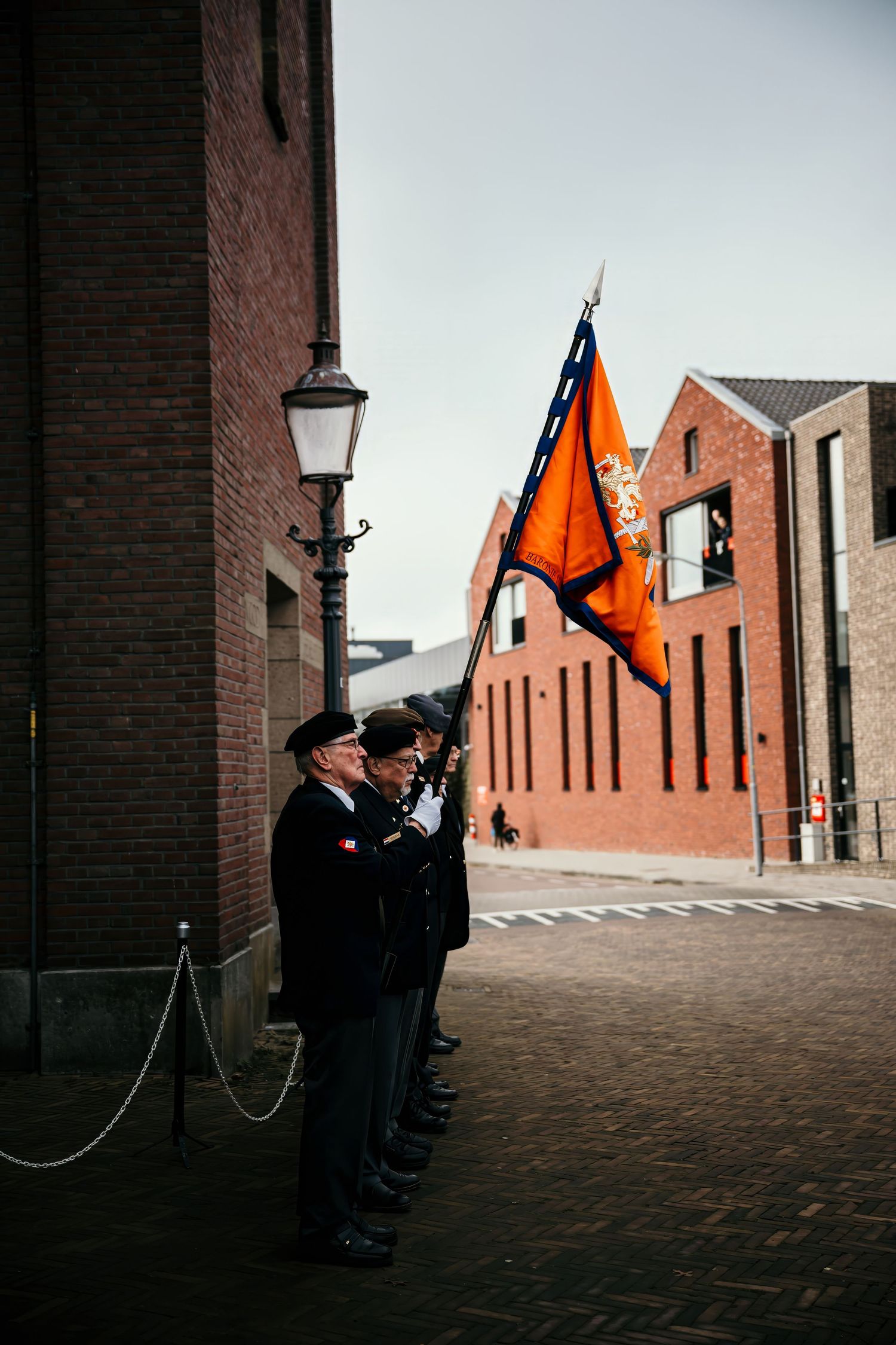 Flag bearers in dark uniforms stand at attention along a brick wall with a traditional street lamp in Liverpool.