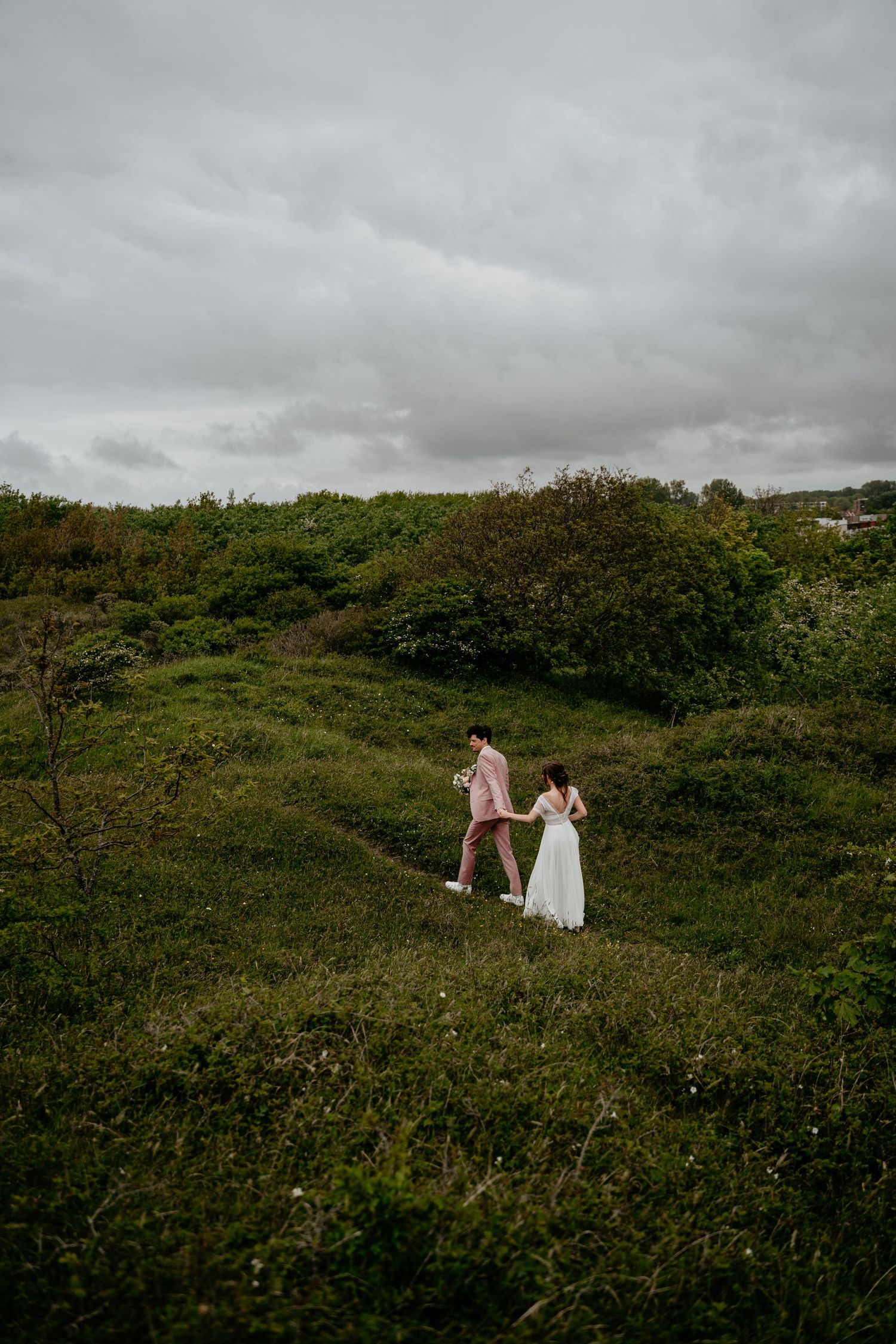 Wedding couple poses for romantic photos on a green hillside under cloudy skies.