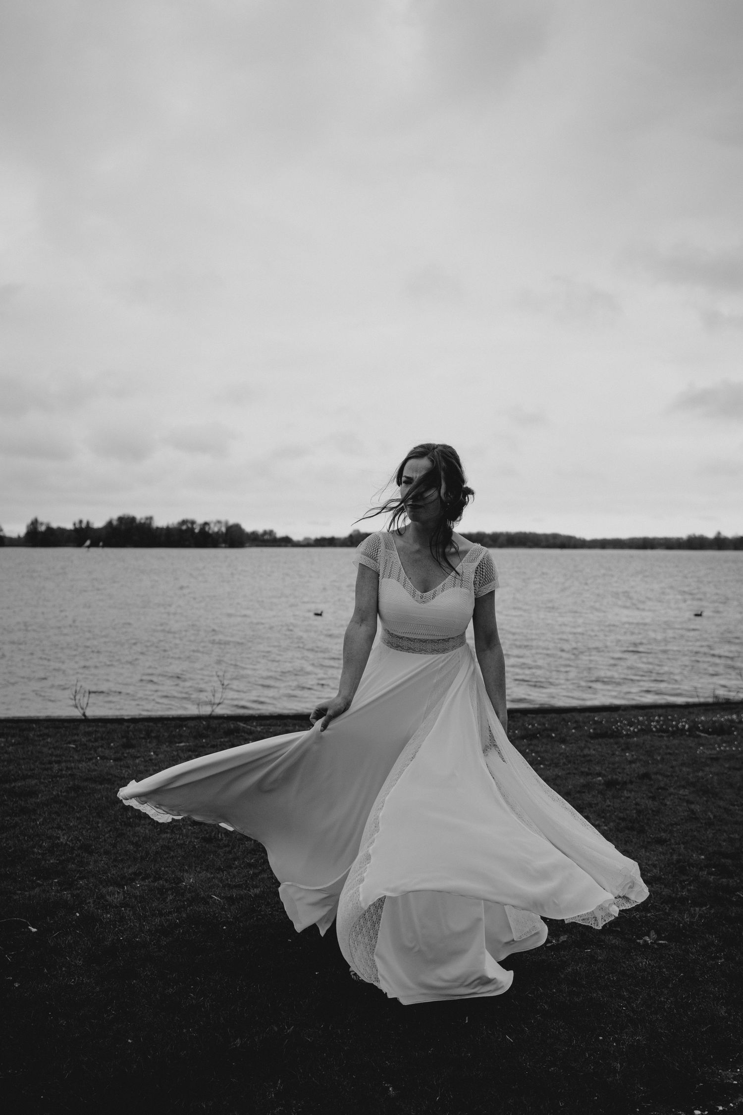 Person in flowing white dress twirling by the waterfront on overcast day.