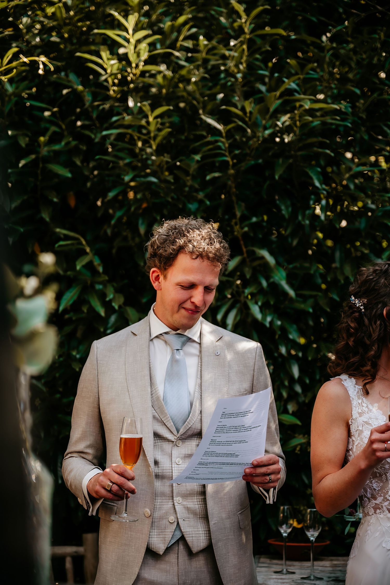 A couple reads wedding vows and holds drinks in a garden setting with lush foliage in the background.