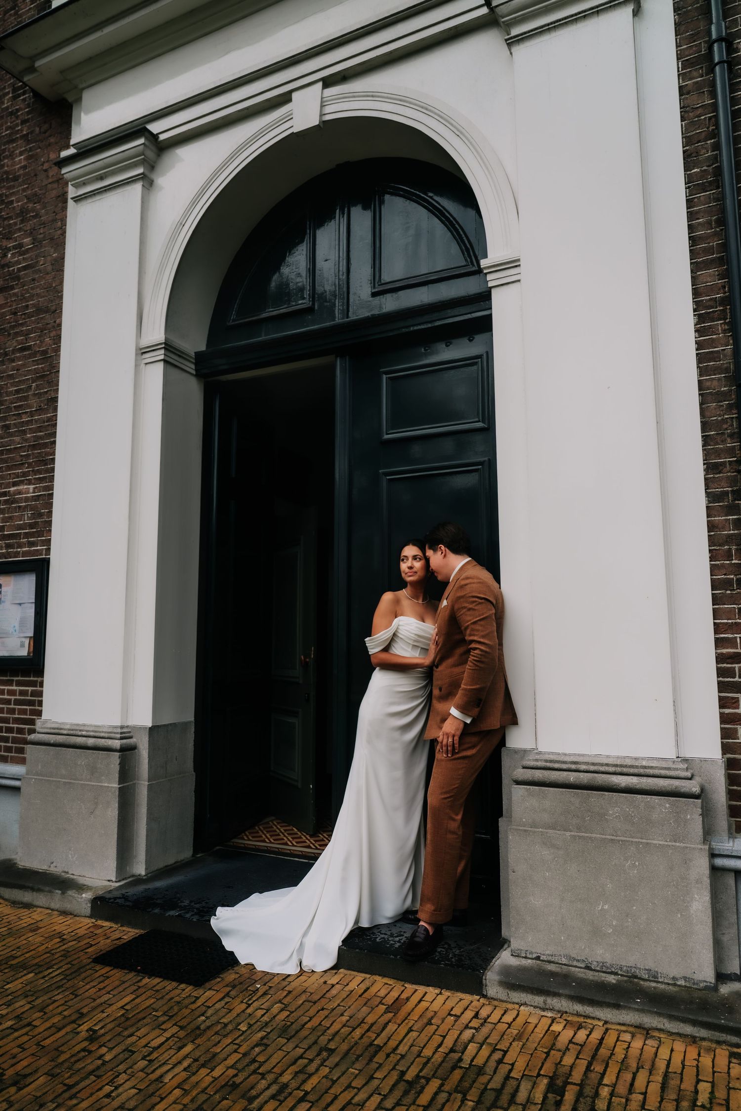 Bride in white gown and groom in brown suit embrace in arched doorway of historic brick building.