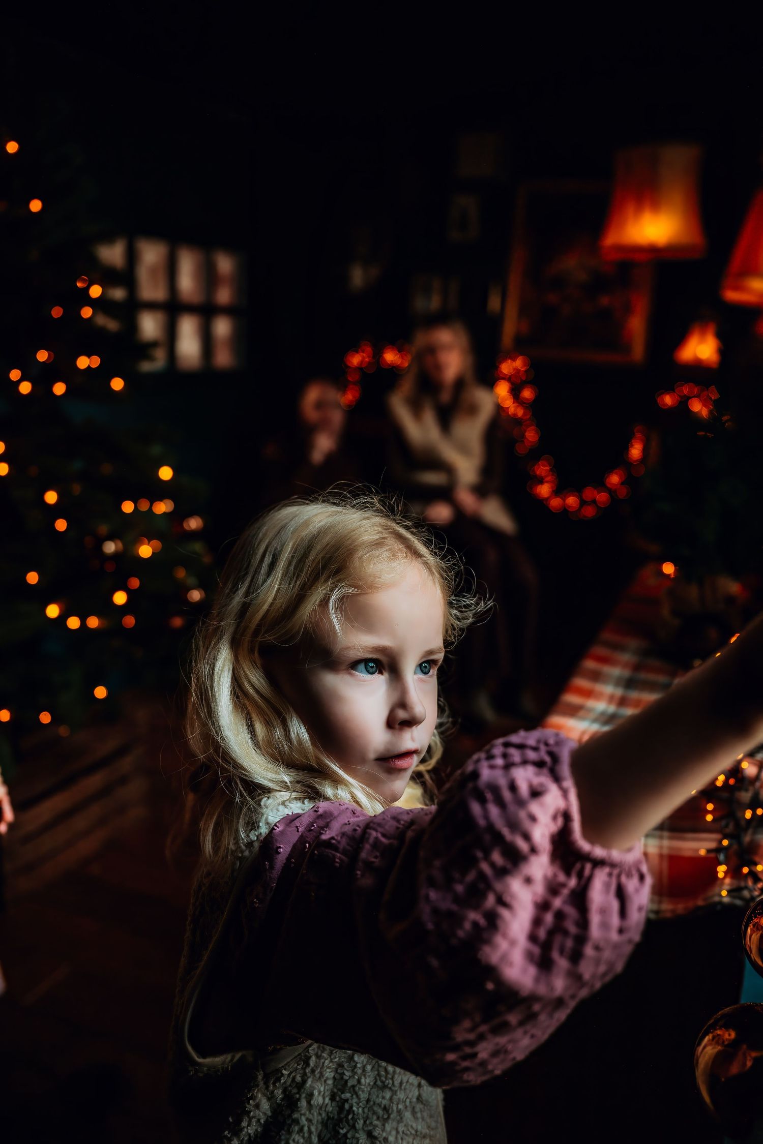 A child in a pink sweater reaches toward twinkling Christmas lights in a dimly lit room.