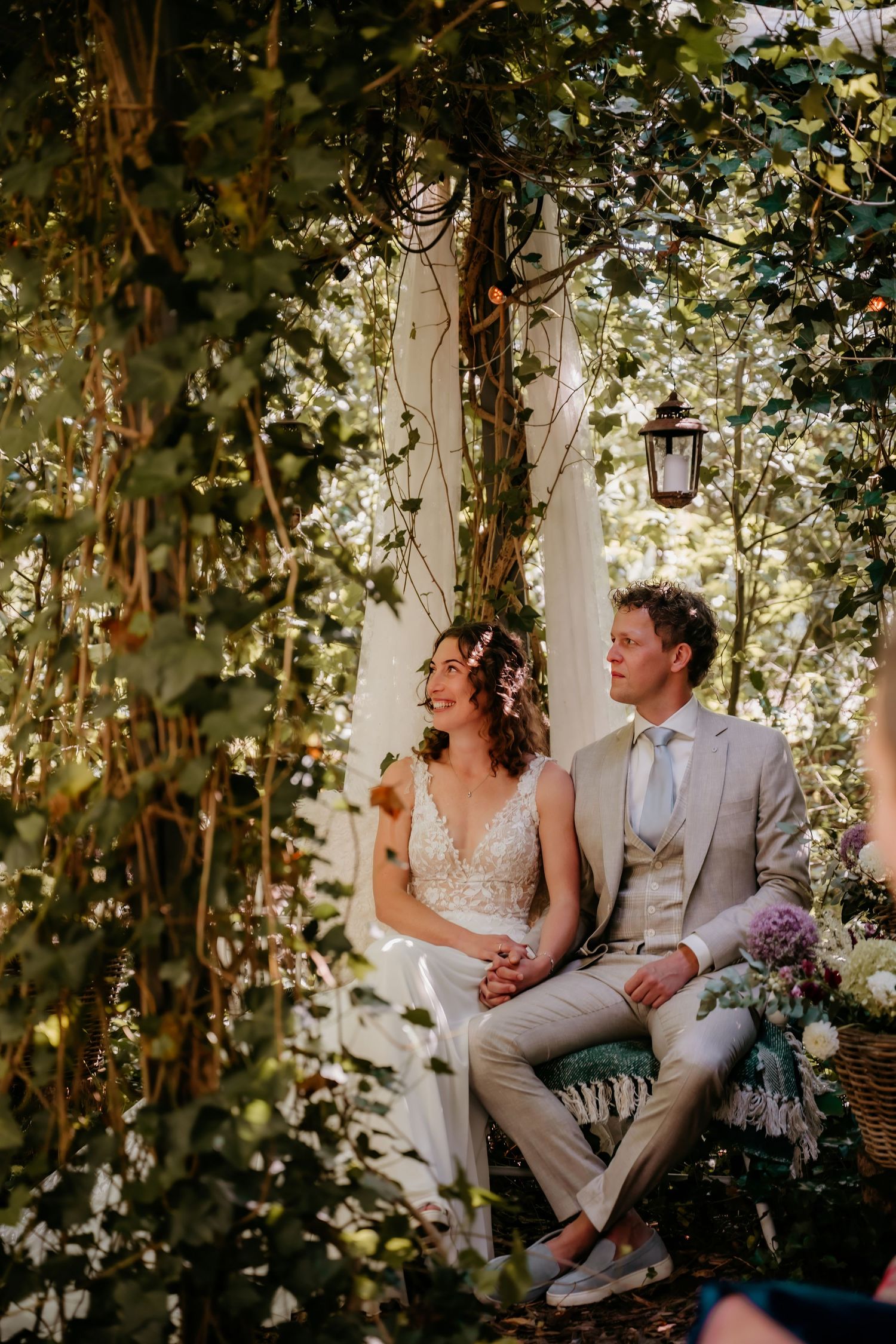 Couple sitting on garden bench surrounded by lush greenery and twinkling lights at outdoor wedding venue.