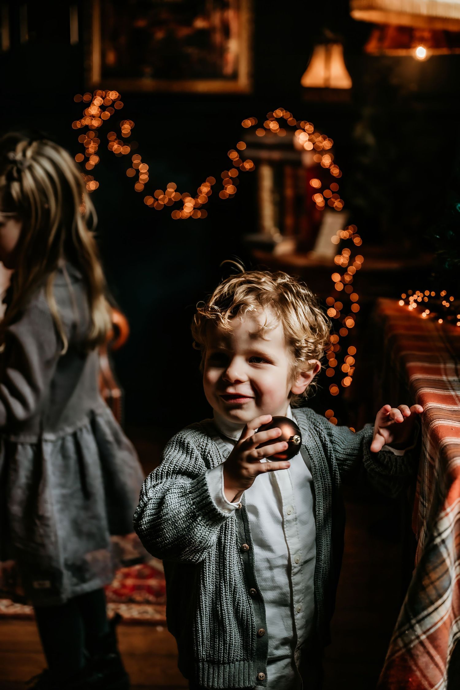 A child in a gray sweater laughs and plays in a dimly lit room decorated with warm glowing string lights.