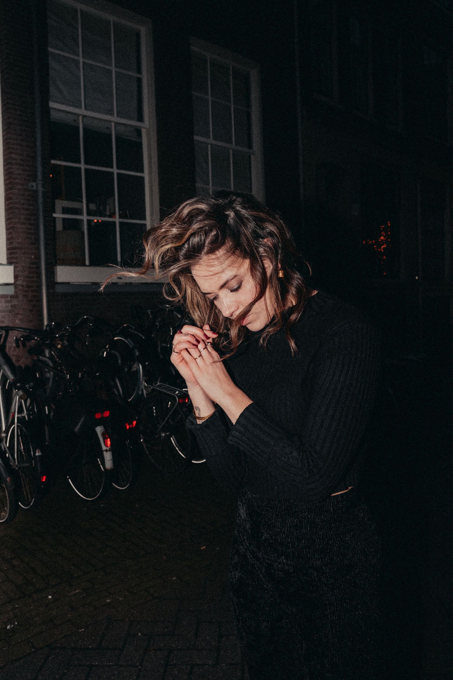 A series of dark moody photographs showing a person with long hair crouching near parked bicycles at night.
