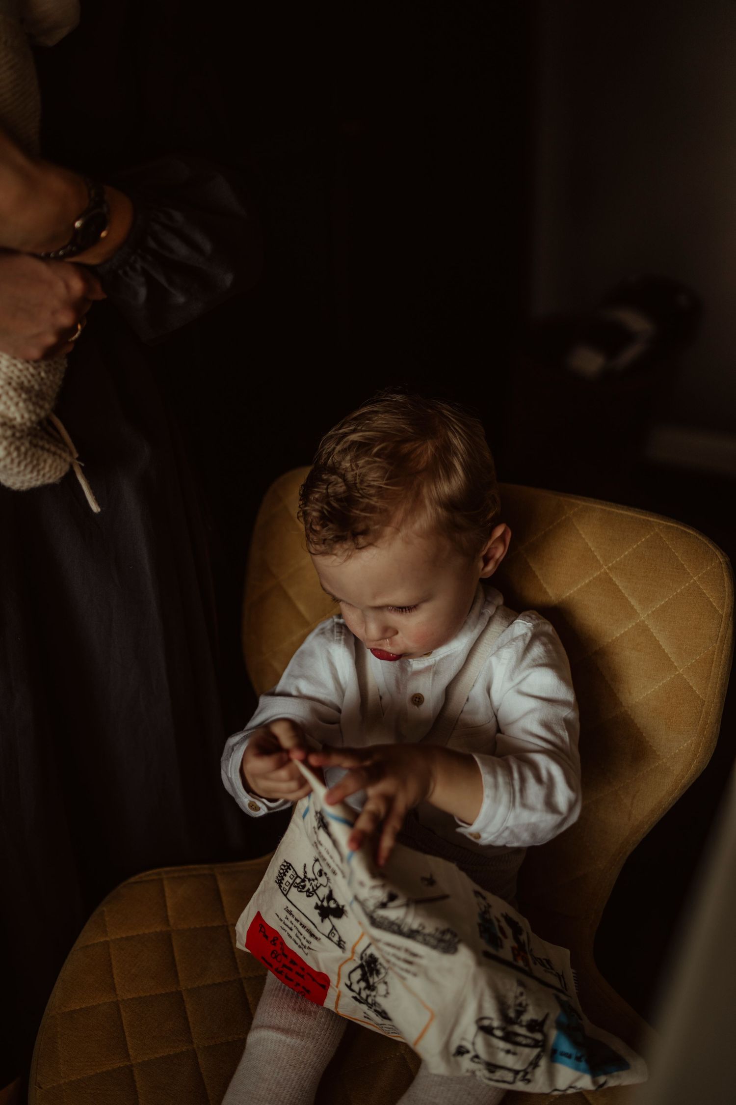 A small child in white clothing sits in a quilted chair examining something intently.
