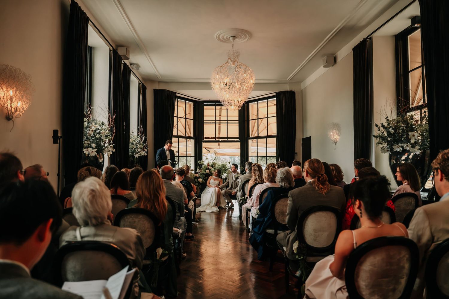 Elegant wedding ceremony in a grand room with chandelier, guests seated in rows facing the couple at the altar.