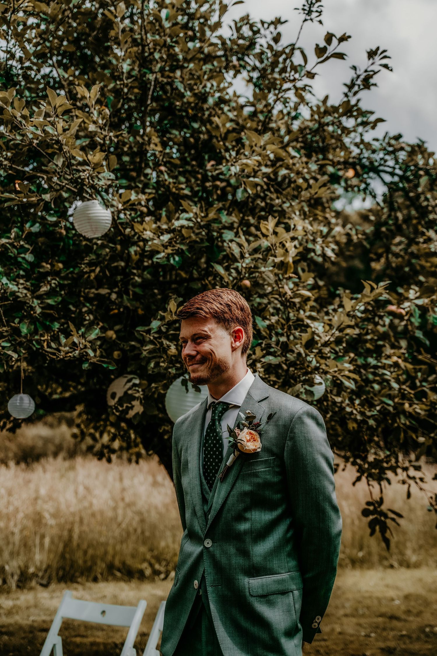A groom in a dark suit stands in front of lush greenery and hanging lanterns at an outdoor wedding venue.