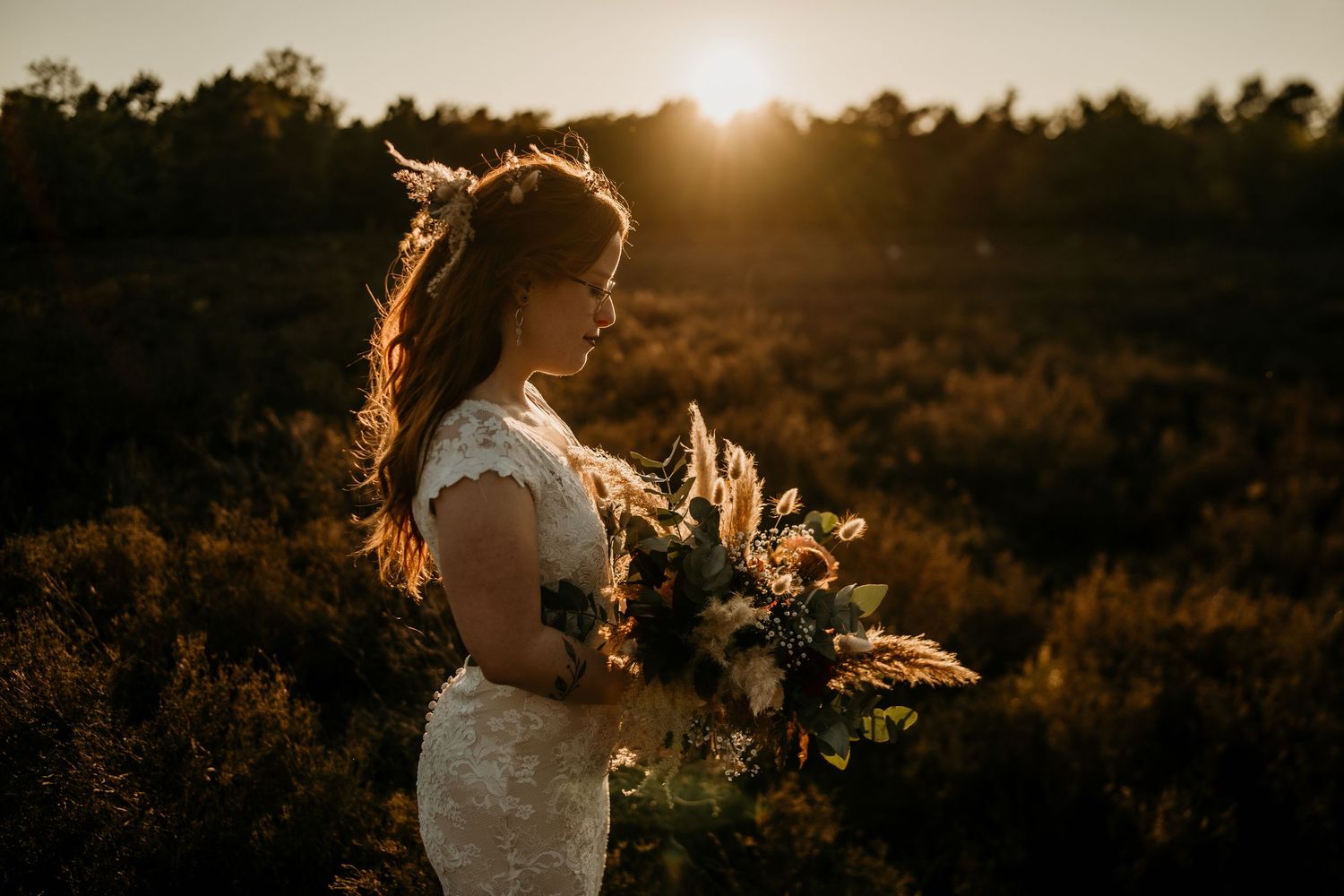 Bride holding bouquet in golden sunset light with flowing dress and natural backdrop.