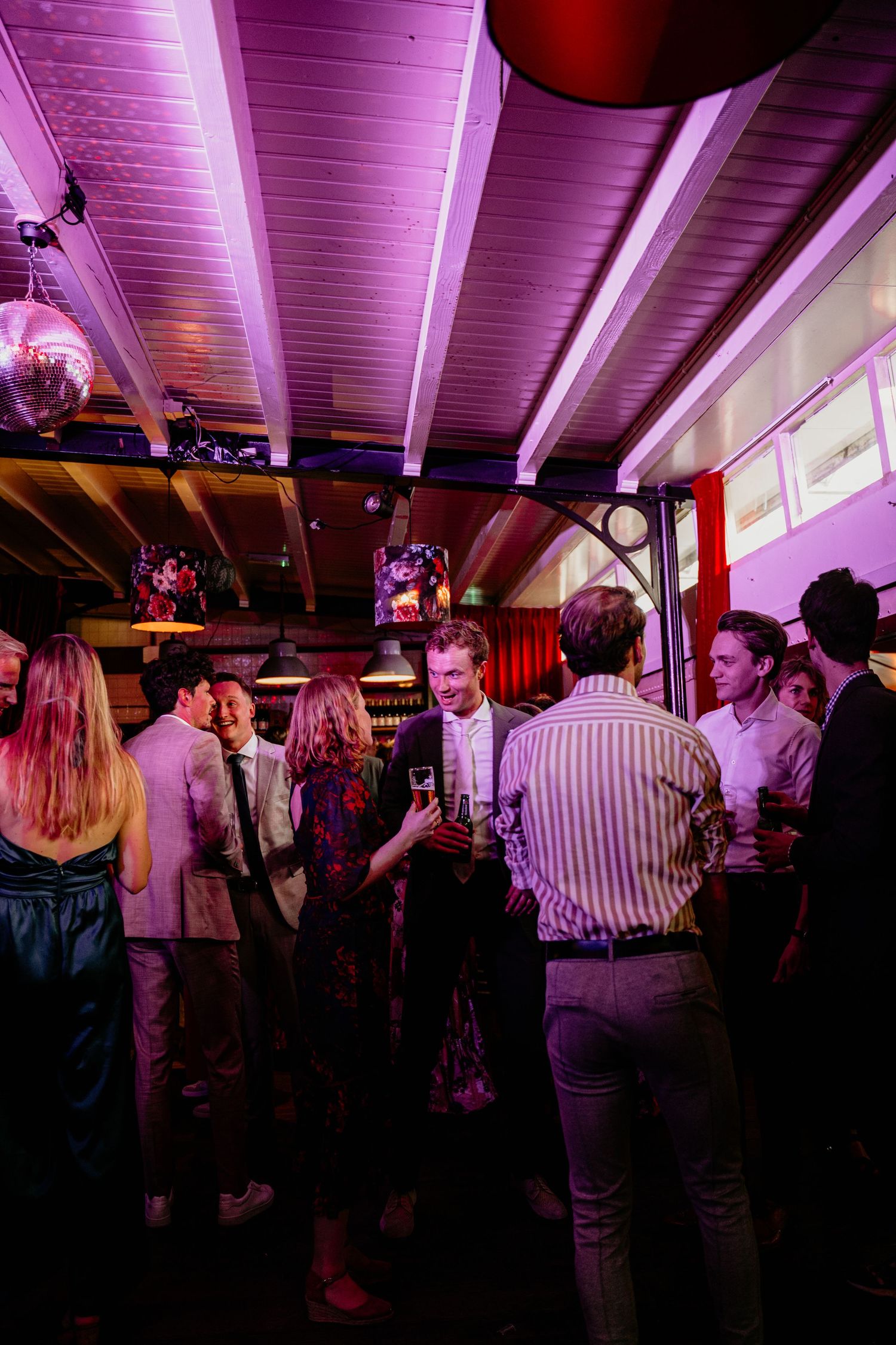 Party guests mingling under purple-lit ceiling beams in a dimly lit venue space.