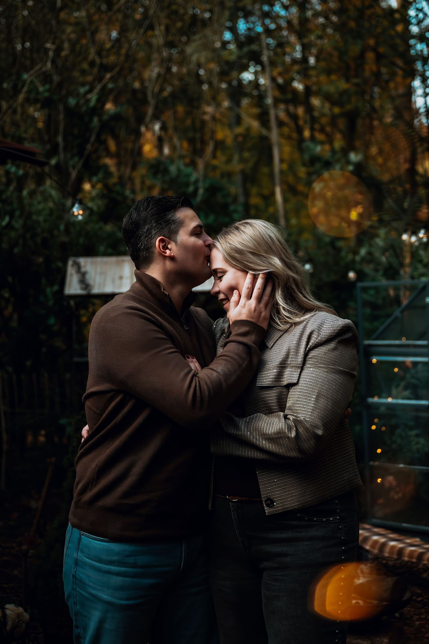 A couple shares a romantic moment on a park bench with autumn foliage and a pumpkin in the background.